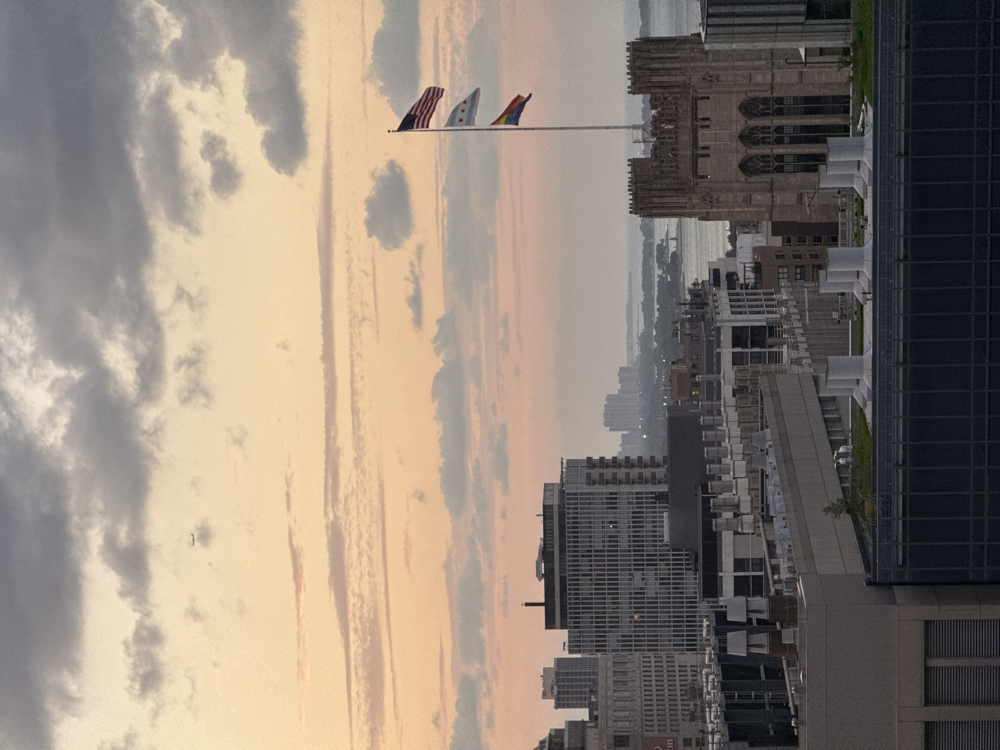 The image captures a cityscape of Chicago, USA, at dusk. The sky is filled with soft orange and pink hues, interspersed with gray clouds. In the distance, a body of water, likely Lake Michigan, is visible, with a faint outline of buildings on the horizon.
In the foreground, several buildings of varying architectural styles are prominent. To the right, a large, gothic-style building with arched windows stands tall. A flagpole is mounted on top of this structure, flying three flags: the American flag, the Chicago city flag, and a rainbow pride flag. To the left of this building, a cluster of modern high-rise buildings with numerous windows dominates the scene.
The rooftops of several buildings are visible, featuring HVAC equipment such as vents and chimneys. A small airplane is spotted in the upper left portion of the sky. The overall impression is a panoramic view of urban development against a tranquil, dusky sky.
