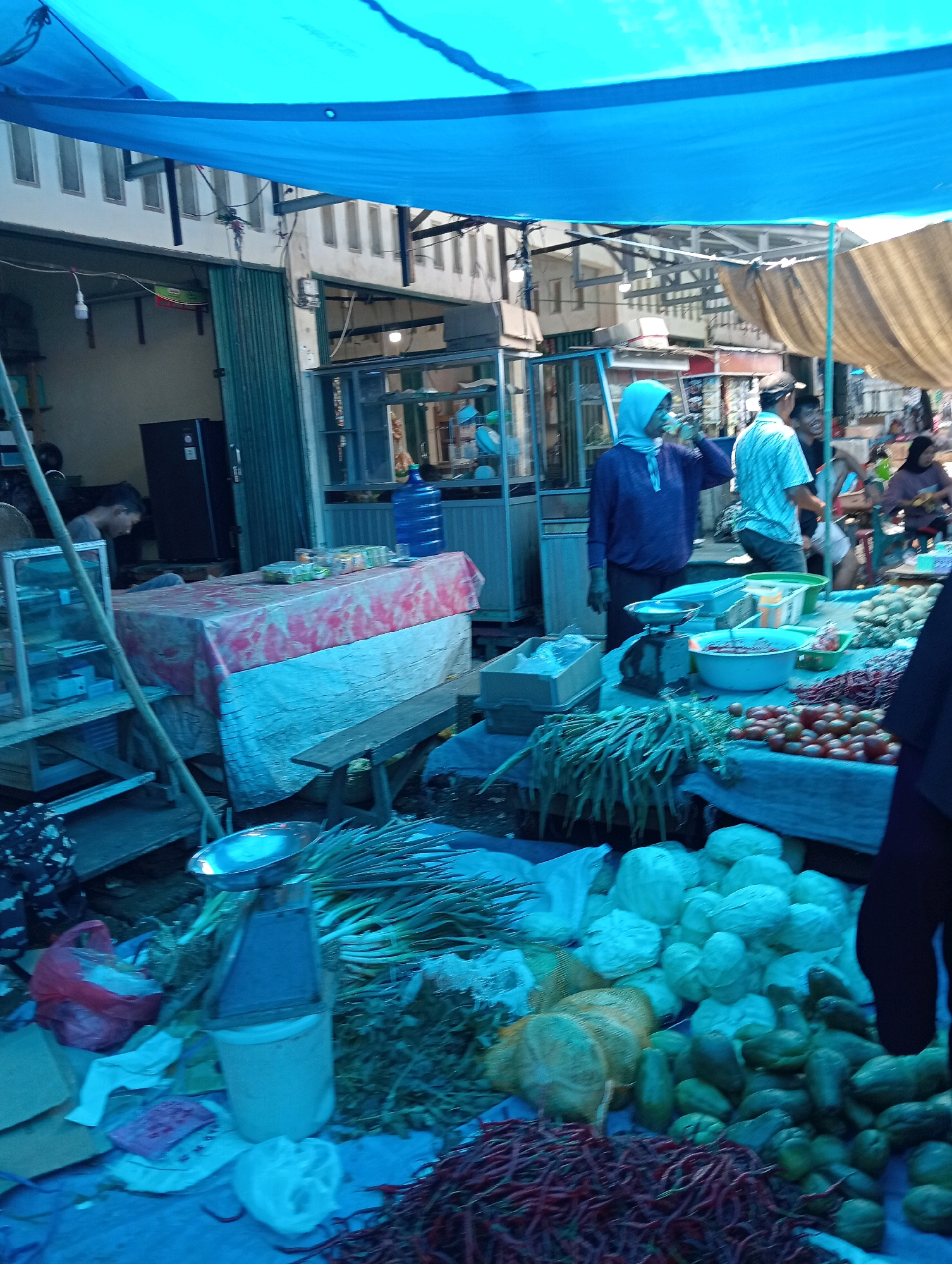 The image depicts an outdoor market scene in Pariaman, Indonesia, likely a bustling local market. A large blue tarp is stretched overhead, providing shade. In the foreground, various fresh produce is displayed on a blue cloth-covered surface. There are bundles of green beans, piles of tomatoes, and what appear to be chiles and cabbages. A vintage scale is positioned on a bucket, suggesting it's used for weighing produce. Further back, a table is covered with a red and white patterned cloth, possibly displaying more goods or acting as a counter. Several people are visible in the market. A woman wearing a blue hijab is standing near the table, and other individuals, including men in casual shirts, are seen browsing or working. Behind the stalls, there are buildings with corrugated metal and tiled roofs, characteristic of local Indonesian architecture. The overall impression is of a lively, open-air marketplace with a focus on fresh food sales.