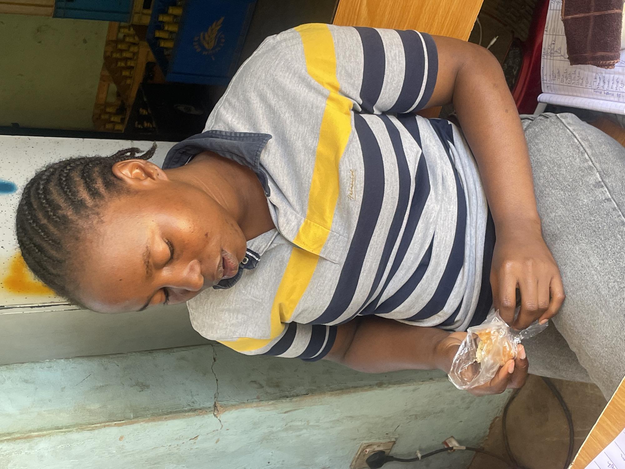 A person is seated at a table in what appears to be a shop in Jos, Nigeria. The individual is wearing a striped, short-sleeved polo shirt with a yellow stripe and light grey jeans. They have dark skin and braided hair. They are holding a plastic bag containing what appears to be a piece of food. The background includes a blue crate, a notebook, and a light blue wall. The scene is lit by natural light. There are some electrical wires and a power outlet visible beneath the table.