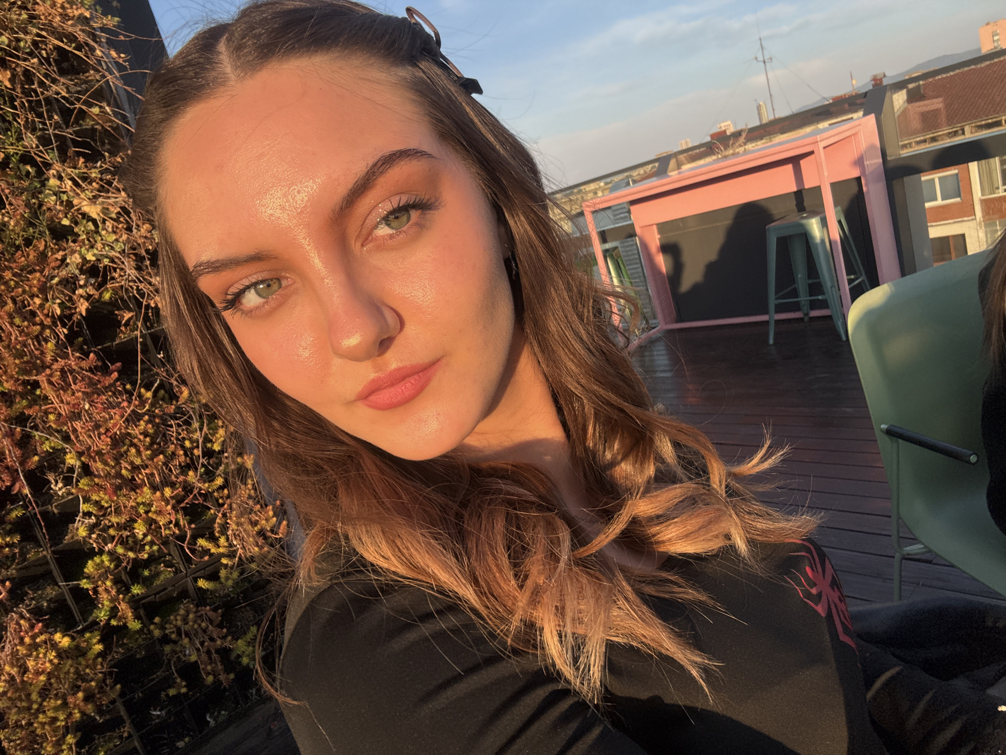 A close-up selfie of a young woman with long brown hair, parted in the middle and styled with a black hair clip, is taken outdoors on a sunny day in Sofia, Bulgaria. Her skin is glowing, possibly from the sunlight. She has fair skin, green eyes, and defined eyebrows. Her lips appear to have lipstick applied.
Behind her, a wall of foliage is visible. In the background, there's a pink structure with black detailing and a teal-colored stool nearby, suggesting a rooftop bar or lounge area. The broader cityscape of Sofia is partially visible beyond the pink structure, including buildings and rooftops.
The woman is wearing a black shirt with a red design visible on the sleeve. The lighting suggests the photo was taken during the late afternoon or early evening when the sun is low in the sky.