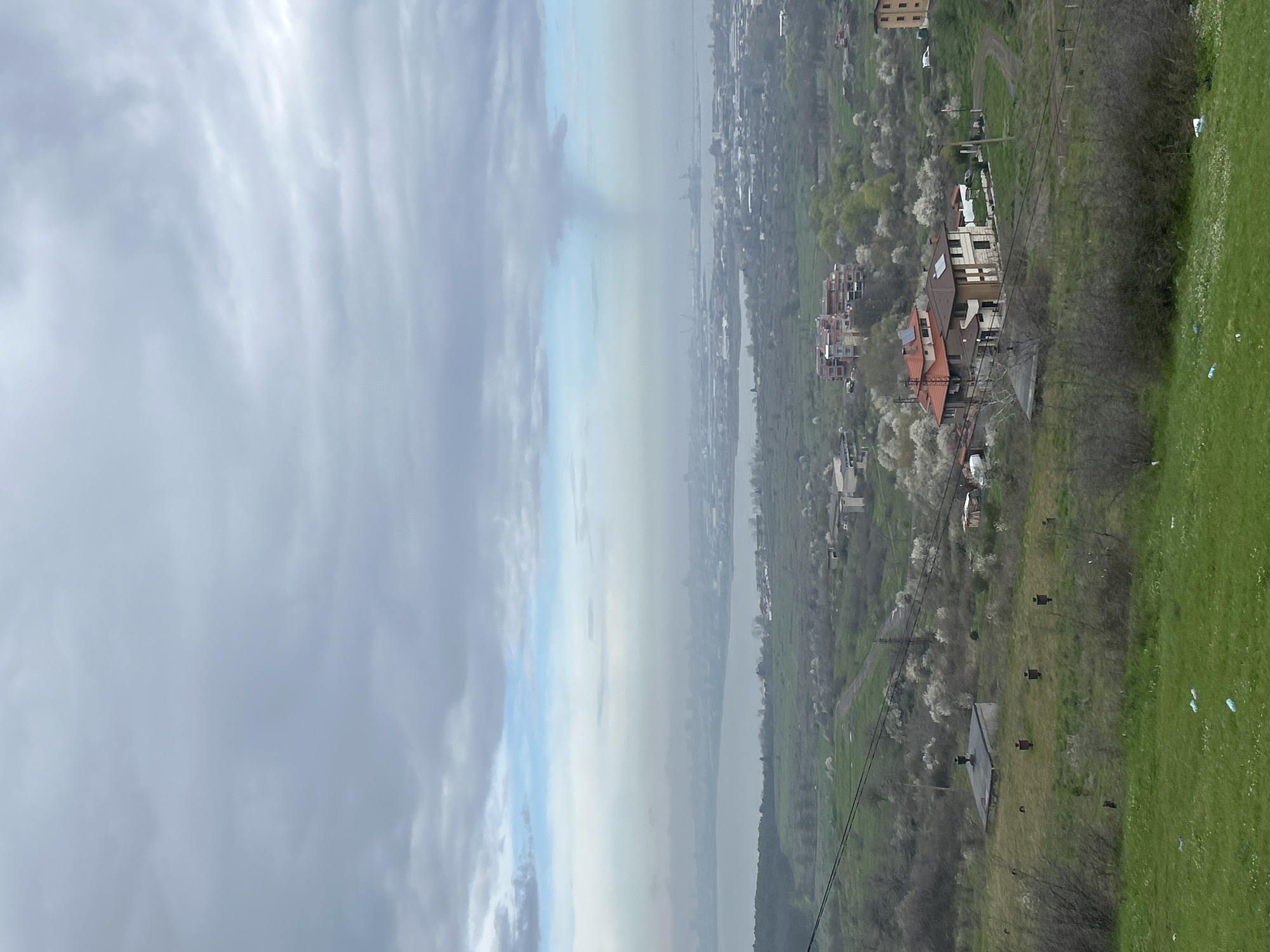 The image depicts an elevated view of a landscape under a cloudy sky. On the right side of the image, green vegetation and buildings are visible. The land slopes downward, with a mixture of grass and trees. Some buildings have red-tiled roofs. The sky is predominantly gray, with patches of light blue. In the center, a body of water stretches towards the horizon. The opposite shore features a city with structures visible in the distance. The city's buildings appear dense. The image is taken from a high vantage point, potentially a hill or tall structure. While the presence of Burgas, Bulgaria is suggested, it cannot be forensically confirmed from this image alone. No people are clearly visible. The dominant actions are the presence of the landscape and cityscape.