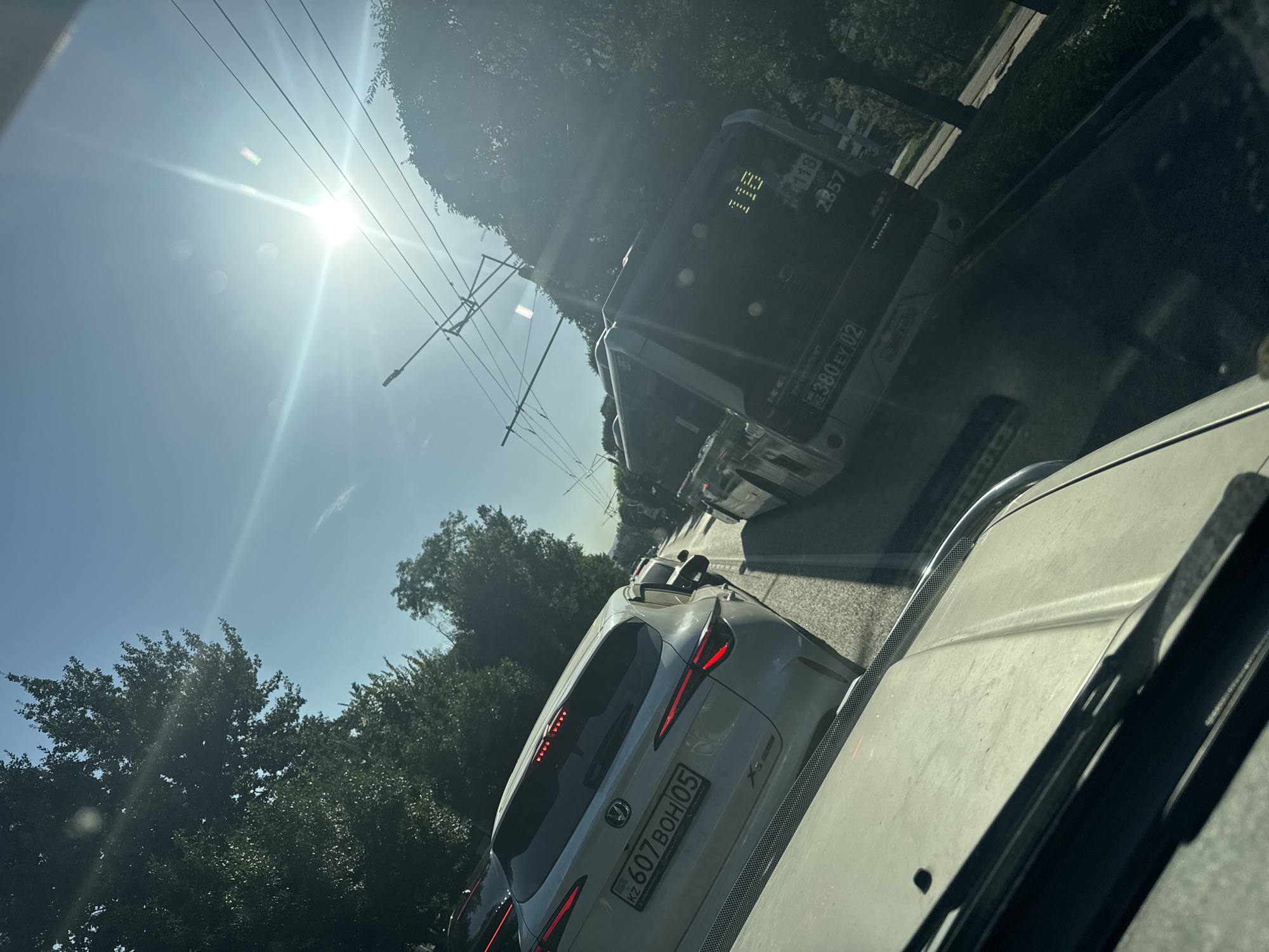 The image captures a street scene in Almaty, Kazakhstan, during the daytime, with a bright sun in the sky creating lens flare. The perspective is from inside a vehicle, looking forward. A white SUV with license plate "KZ 607 BOH 05" is in the foreground on the left. To its right, a dark-colored bus is visible, its rear displaying route number "118" and vehicle number "2857". The bus's license plate is partially obscured but appears to start with "KZ 380 EY 02". Overhead, trolleybus wires are strung across the street, indicating public transportation infrastructure. Lush green trees line the street on both sides, suggesting an urban area with significant vegetation. The traffic appears to be moderate, with other vehicles visible in the distance. The overall impression is a typical urban commute in Almaty on a sunny day.