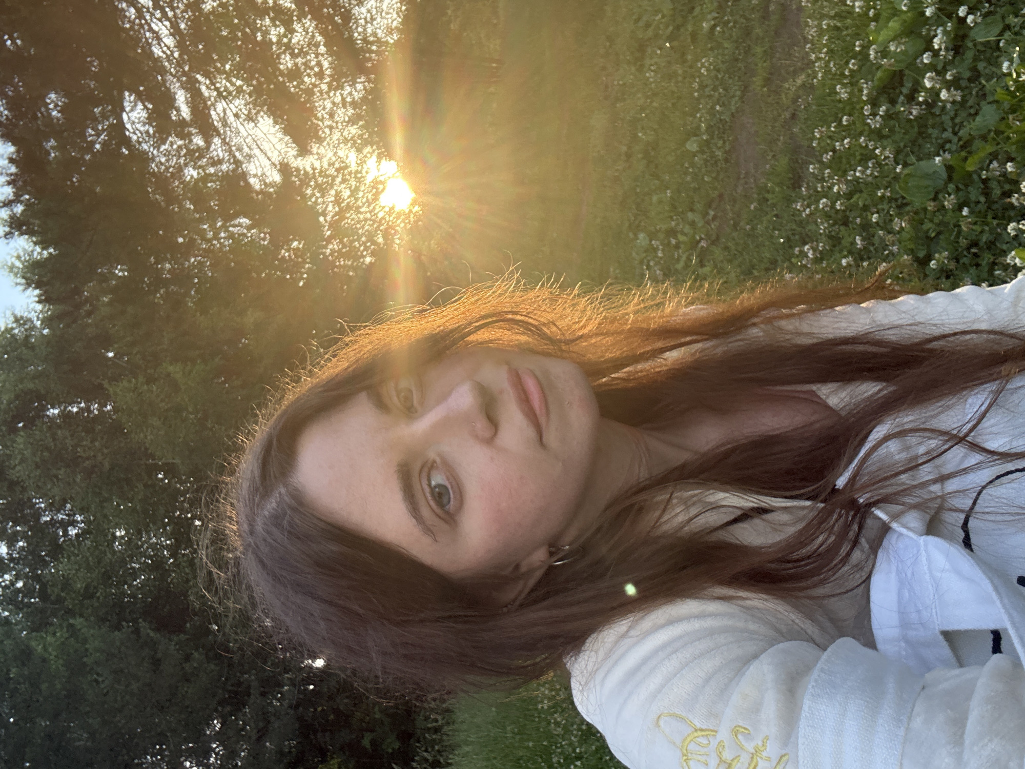 A close-up, high-angle medium shot shows a young woman with long, dark brown hair in the foreground, facing slightly to the right and looking directly at the camera. She is wearing a light-colored jacket with yellow embroidery. The sun is setting behind her, casting a warm, golden light and lens flare across the scene. The background consists of green grass and trees, with a few small white flowers scattered in the grass. The image appears to be taken outdoors during the golden hour. The specific location within the city of Unknown, Unknown, is not discernible from the image itself.