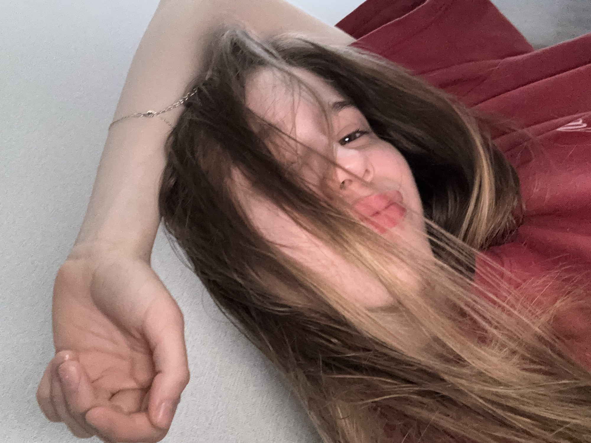 A young woman is captured in a close-up, selfie-style photograph. She has long, layered brown hair with lighter streaks, which partially obscures her face. Her lips are slightly parted, and she appears to be looking towards the camera with a playful or seductive expression. Her right arm is raised and bent, with her hand resting near the back of her head. She is wearing a dark red t-shirt. A delicate silver bracelet is visible on her wrist. The background is a plain, light-colored wall. The overall lighting suggests an indoor setting with soft illumination. There are no discernible elements in the image that would indicate a specific location in Sinyavino, Russia.