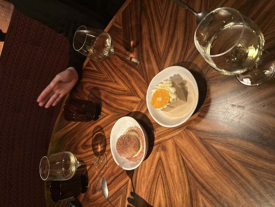 A top-down view showcases a textured wooden table. Two white plates, one with a dessert featuring a slice of orange and whipped cream and the other with a cocoa-dusted dessert, sit between two wine glasses and water glasses. A hand extends onto the table near one of the wine glasses. Silverware is also present near one of the dessert plates.