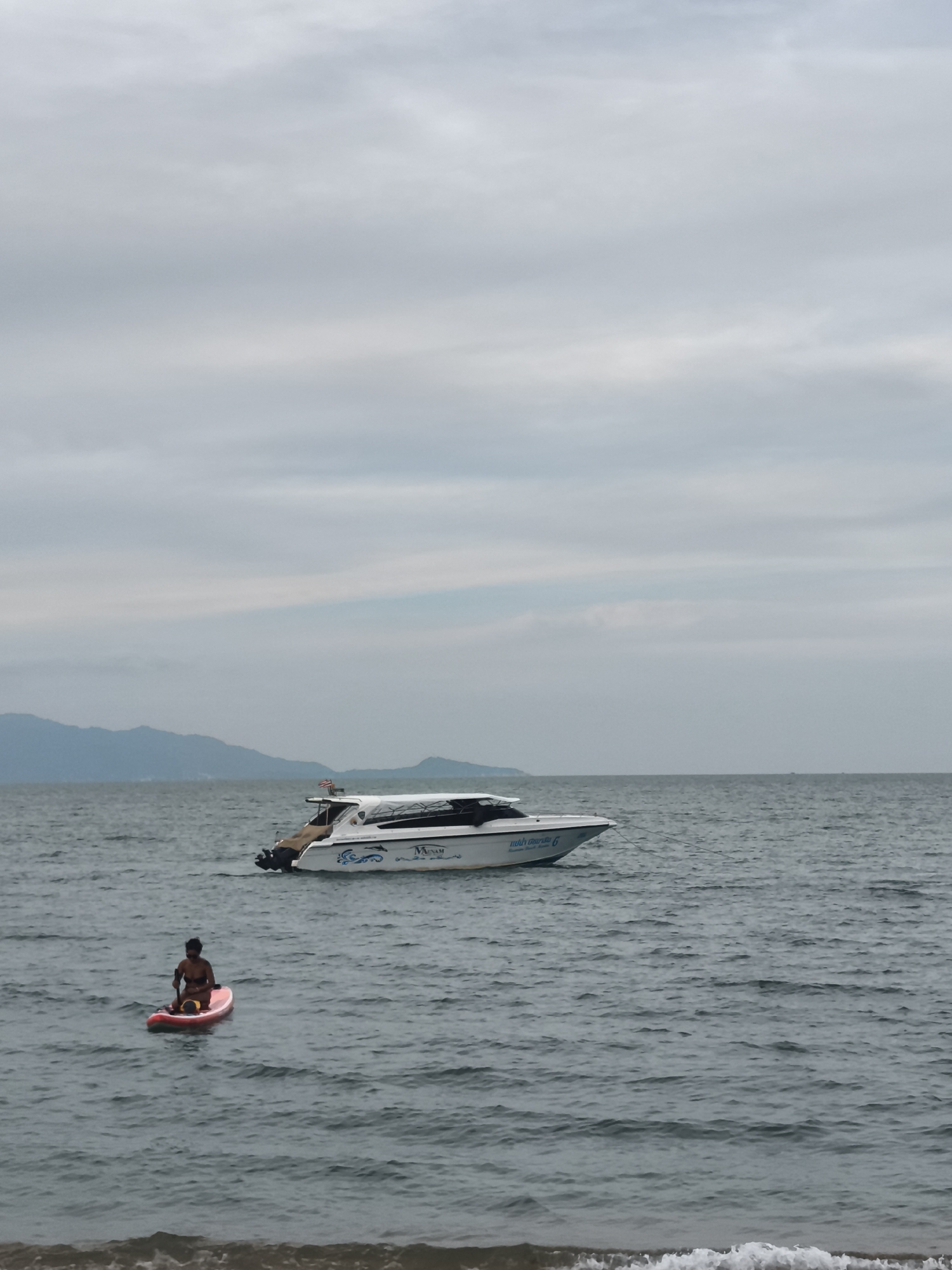 The image depicts a tranquil coastal scene in Baan Mae Nam, Thailand, under an overcast sky. In the foreground, a person is seated on a bright red paddleboard, facing away from the camera, holding a paddle. The person appears to be wearing dark swimwear. Gentle waves are visible along what seems to be the edge of a sandy beach in the bottom right corner.
Further out in the dark grey-blue sea, a white speed boat with two outboard motors is visible. The boat features blue wave-like graphics on its hull. Prominently displayed text on the boat reads "MNNAM Samui Speed Boat" and includes Thai script, specifically "แม่น้ำ ชอย 6" (Mae Nam Soi 6), along with the number "6". An American flag is flying from the stern of the boat.
In the distance, a range of mountains extends across the horizon, silhouetted against the uniformly cloudy sky. The lighting is soft and diffused, indicating either an overcast day or possibly early morning/late afternoon. The overall scene suggests a calm recreational setting, with the person paddleboarding and the boat idling in the water.