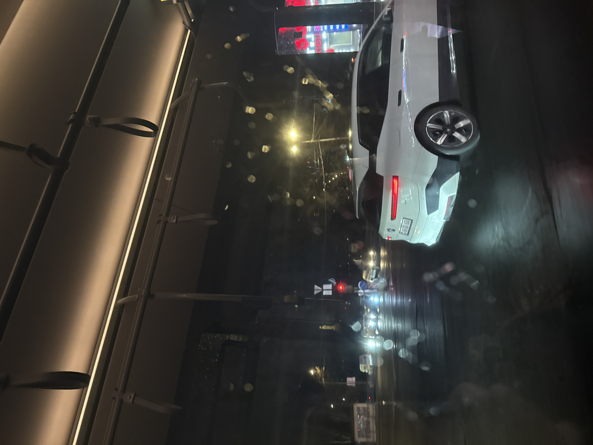 The image shows a nighttime street scene, likely taken from inside a bus or train. The viewer looks out through a rain-splattered window. Reflections on the glass add to the blurred effect. A white car is visible on the right, illuminated by streetlights and its own rear lights. In the background, blurred lights suggest a city street with other vehicles and signs. The interior of the bus or train is partially visible on the left, showing overhead bars for passengers to hold on to. Given the urban setting and the type of vehicle, it is possible this scene is in Yerevan, Armenia. However, without additional context or visual confirmation, it is impossible to conclusively confirm the location.