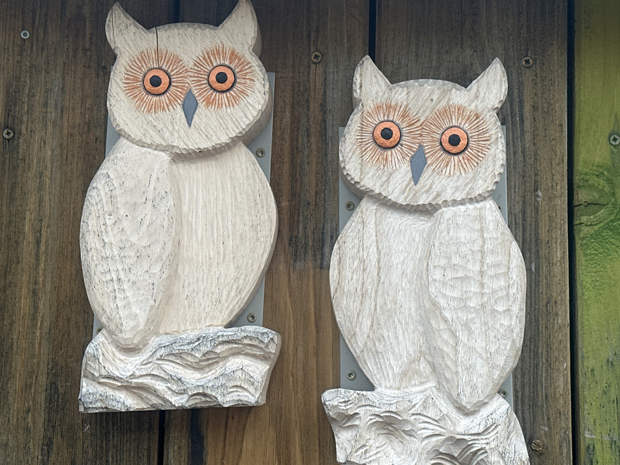 Two wooden owl carvings are mounted vertically on a weathered wooden fence. The owls are carved from light-colored wood and feature large, round orange eyes with black pupils and a gray diamond-shaped beak. They have pointed ear tufts and textured bodies. Both owls are perched on carved bases that resemble rocky terrain. The left owl is slightly closer to the camera than the right owl. The fence is made of vertical wooden planks with visible grain and some signs of age and wear. A green-painted wooden surface is visible on the far right edge of the image. The scene is outdoors, with natural light.