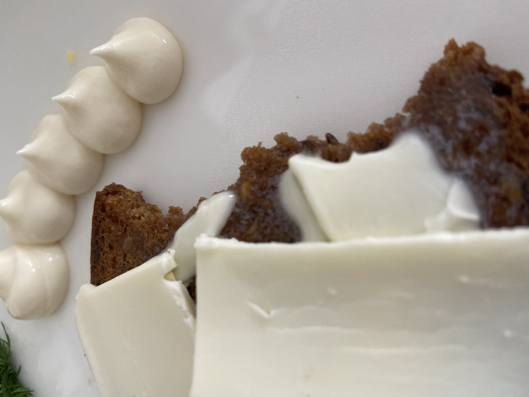 A close-up shot shows a plate of food featuring dark brown bread topped with large, roughly cut pieces of white cheese and dollops of white cream on the side. A sprig of green dill is also visible on the bottom left. The photograph does not provide sufficient information to determine the location in Budva, Montenegro.
