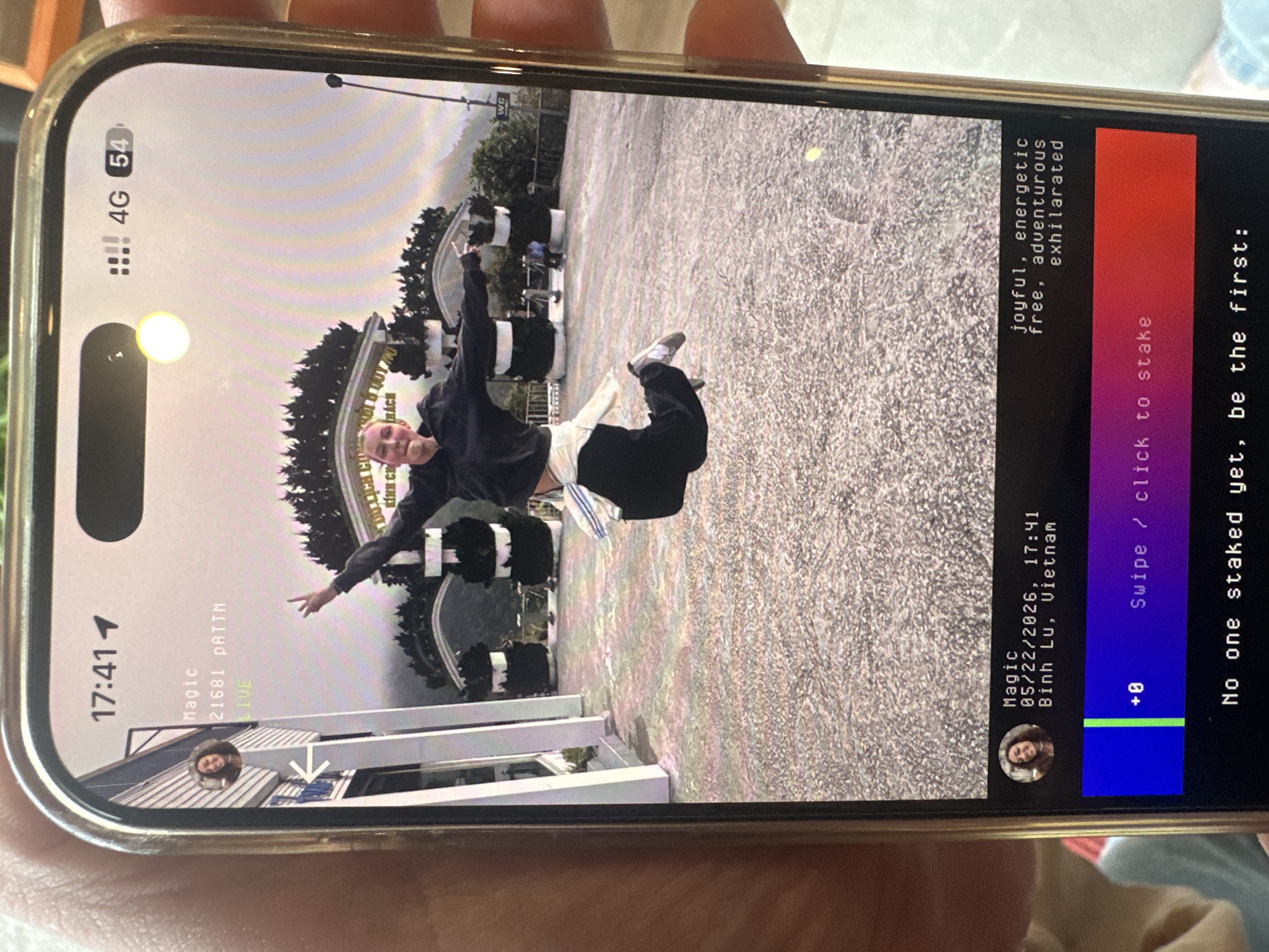 The image shows a cell phone displaying a video or photo of a person in Binh Lu, Vietnam. The person is in front of a sign written in Vietnamese. The phone screen indicates the date as May 22, 2026, and the time as 17:41. There are UI elements of an application, including "Swipe / click to stake," "+0", and descriptors such as "joyful, energetic, free, adventurous, exhilarated". Other information on the top of the phone screen includes "Magic", "21681 PATTN", "LIVE", "4G" and "54". Two small profile pictures are on the screen. The phone is held by a person, whose fingers and hand are visible.
