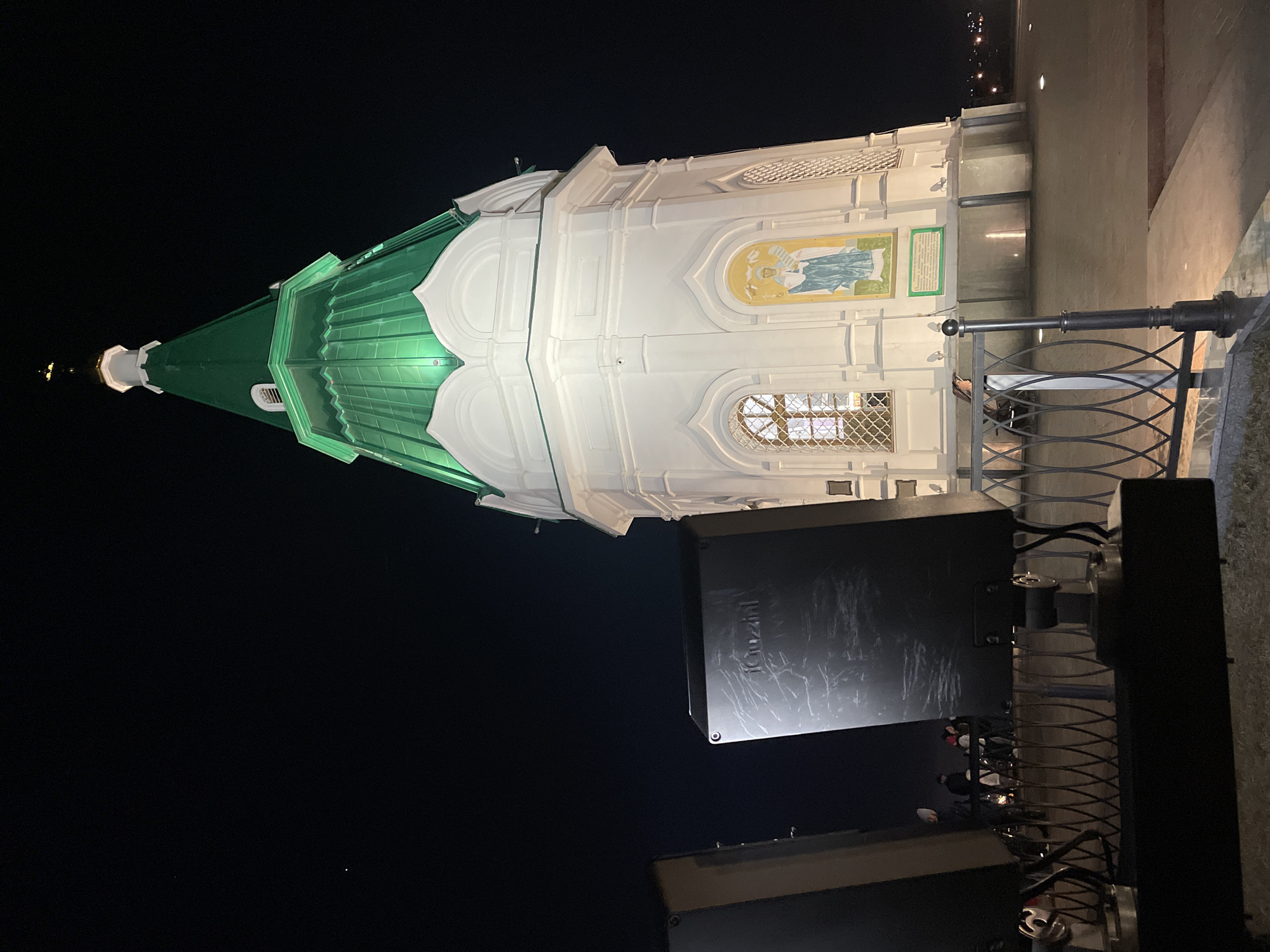 A nighttime view of the Chapel of St. Paraskeva Pyatnitsa in Krasnoyarsk, Russia. The chapel has a green roof and white walls, adorned with religious iconography. The background is mostly dark sky. The chapel sits near a metal fence. There are some electrical boxes nearby.