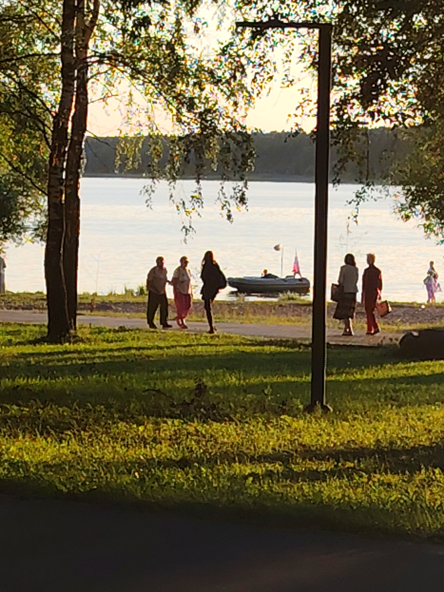 A group of people are walking along a paved path by a body of water, with trees and grass in the foreground and background. The setting appears to be a park or waterfront area in Konakovo, Russia, likely during the daytime with golden hour lighting due to the warm glow on the trees and water.
In the foreground, the image shows a grassy area with dappled sunlight and shadows. A dark, vertical pole, possibly a lamppost, is prominently positioned slightly to the right of the center. On the path in the middle ground, several individuals are visible walking. From left to right, there are two figures walking together, followed by a single figure in a dark outfit, and then two more figures walking in the same direction. Further along the path, near the water's edge, there are at least two more figures. A small inflatable boat with a person in it is moored on the water near a flagpole with a flag. The water stretches across the background, with a distant tree line visible on the far shore. The sky above the water is bright and hazy, suggesting either a sunrise or sunset.