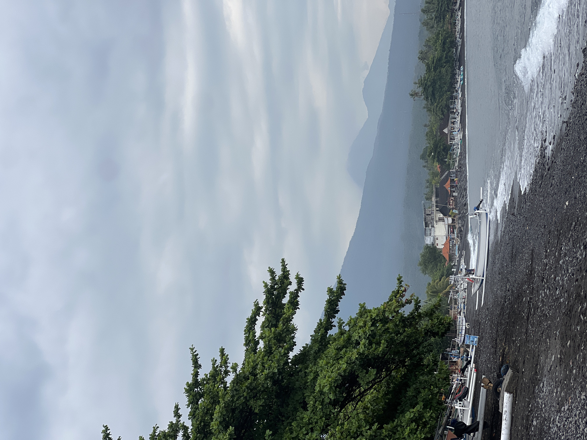 A vertical, eye-level shot captures a tranquil beach scene in Abang, Indonesia, under a cloudy sky. In the foreground, a dark, pebbly shore meets the gentle waves of the ocean. A large, leafy green tree branches out from the left, partially obscuring the view. Several traditional outrigger boats, painted white and with their characteristic outriggers, are lined up on the beach, some partially submerged in the sand. A few individuals are visible near the boats, engaged in what appears to be leisurely activity. Behind the beach, a line of buildings, likely restaurants or accommodations, dots the landscape, with lush green trees and palm trees interspersed. In the distance, a range of misty mountains forms a dramatic backdrop against the muted sky. The overall impression is one of peaceful coexistence between human activity and the natural environment.