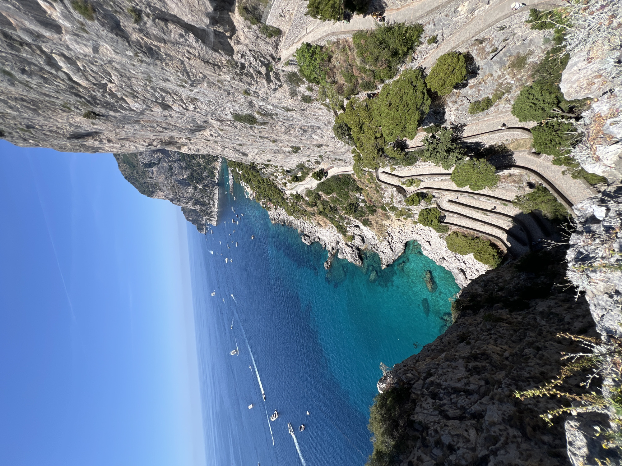 The image depicts a scenic view of the island of Capri, Italy, likely the famous Via Krupp. A steep, winding stone pathway zigzags down a dramatic cliff face. Lush green trees and vegetation cling to the rocky slopes alongside the path, which is lined with stone walls. Below the cliff, the deep blue Mediterranean Sea is visible, dotted with numerous white boats and yachts. The water near the shore is a striking turquoise, indicating its clarity. The sky above is a clear, bright blue, with a thin contrail in the upper left quadrant. The perspective is from a high vantage point, looking down on the winding path and the sea. There are a few people visible on the pathway, appearing as small figures dwarfed by the scale of the natural landscape and the man-made path.