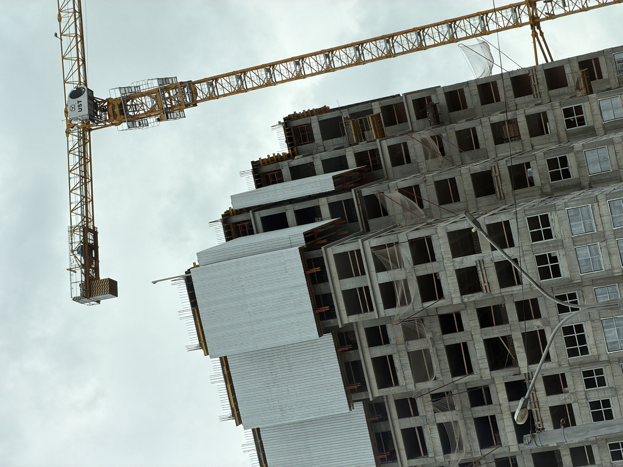 This is a low-angle, outdoor photograph of a large construction site under a cloudy sky, likely in the city of Perm, Russia. A massive, yellow construction crane dominates the right side of the frame, its jib extending horizontally across the upper portion of the image. The crane's main tower is visible vertically, angled slightly away from the viewer. On the left and center of the image, a partially constructed, multi-story building is visible. The building's exterior is made of concrete, with numerous open window frames and rebar protruding from the top of the structure. Large, corrugated metal panels cover sections of the building's upper floors. Two streetlights on curving poles are situated in the lower left corner of the image, obscuring part of the building's base. No people are clearly visible in the scene.