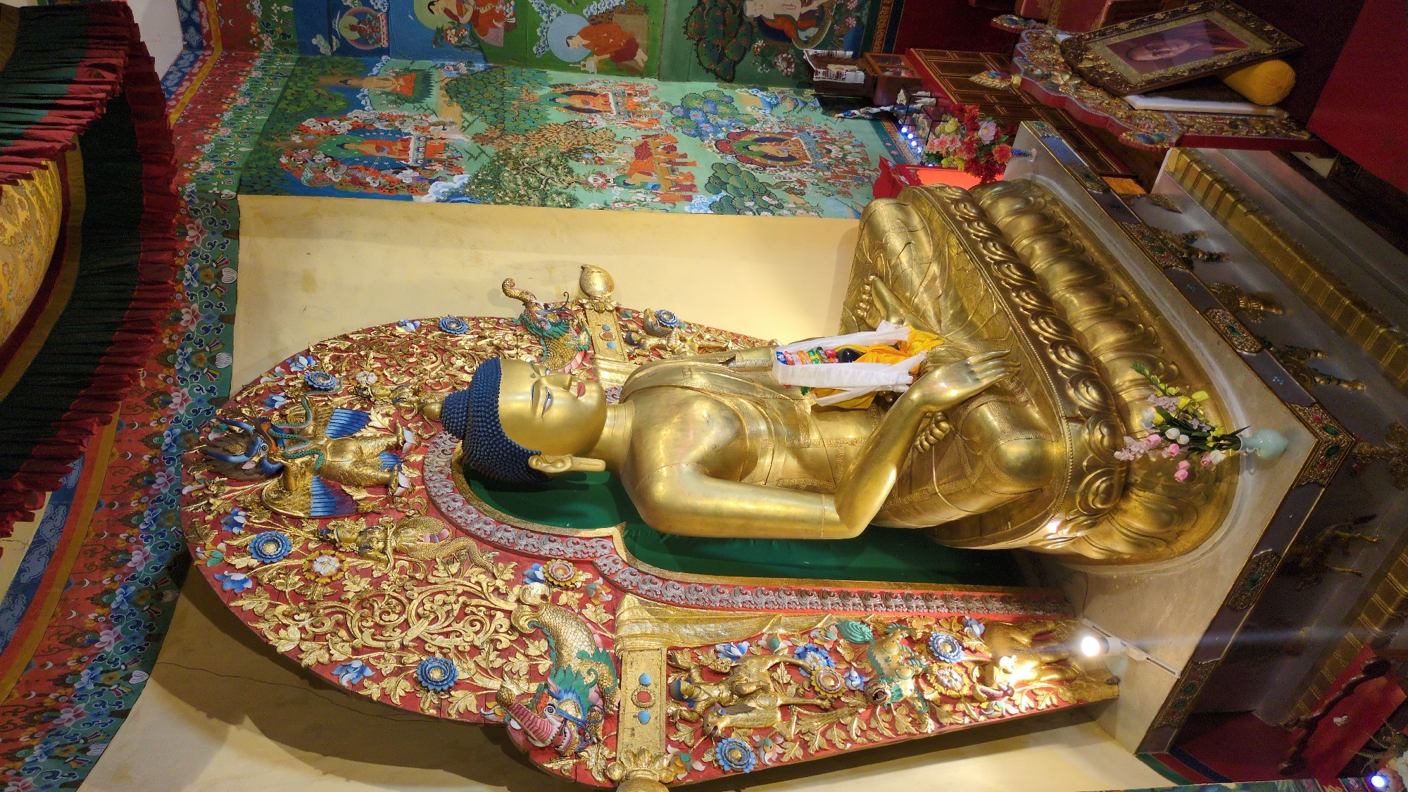 The image depicts a large, ornate, golden reclining Buddha statue. The statue is resting on a red and gold platform adorned with intricate carvings, including depictions of birds. A green fabric cushion supports the Buddha. The statue itself is golden, with a blue headdress and white and yellow scarves placed across its chest. The backdrop includes a yellow wall and a painted mural on the upper portion of the wall and ceiling, featuring various figures and scenes in vibrant colors. To the left, there is a striped fabric hanging. To the right of the statue, there is a display case containing a framed picture and other decorative elements. Based on the presence of a reclining Buddha, the image is likely taken in a Buddhist temple or monastery in Fatehpur, India.