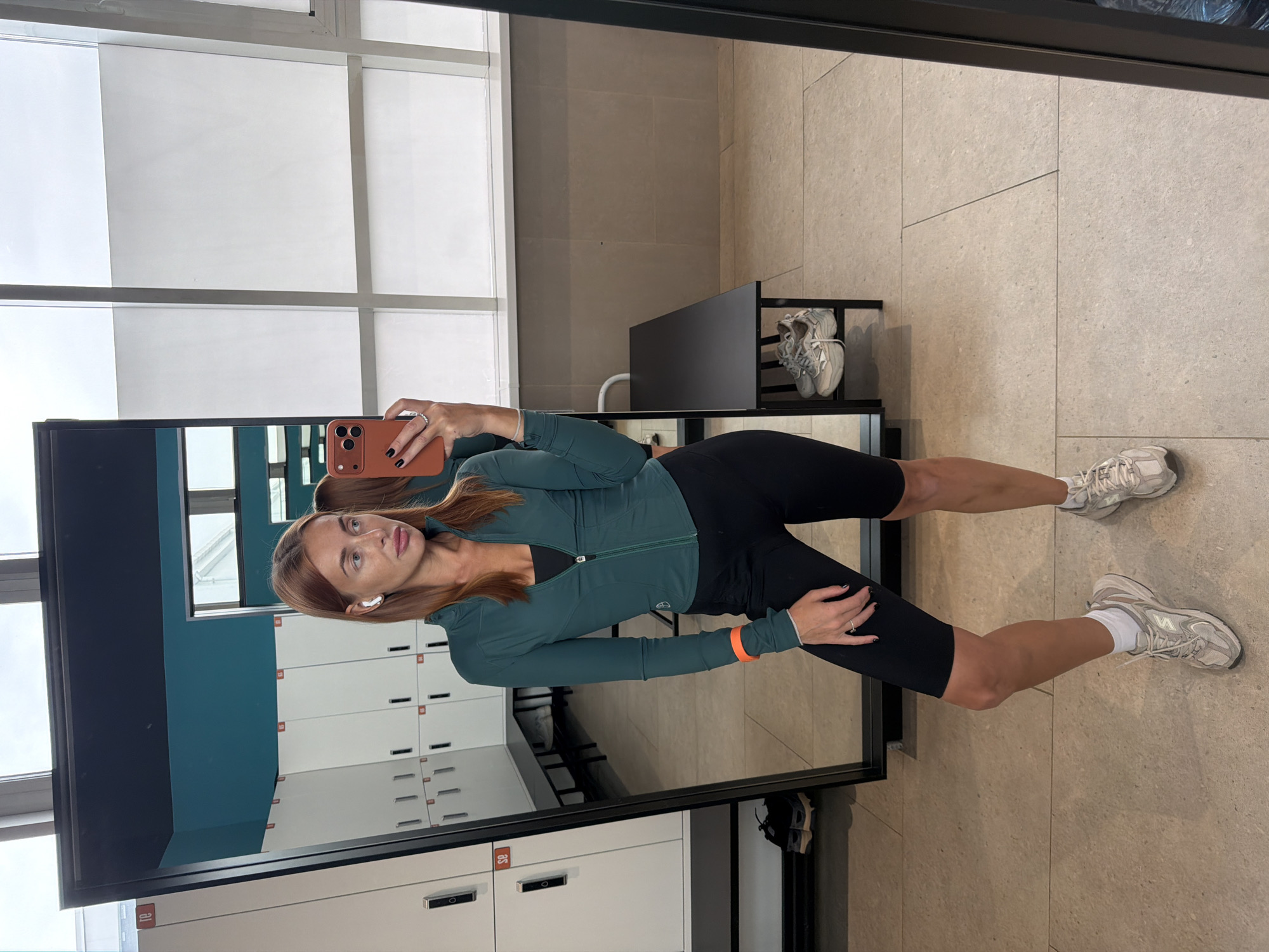A full-body, slightly angled selfie of a woman in athletic wear is taken in a locker room. She has light skin and reddish-brown hair, which is pulled back. She is wearing a dark teal zip-up jacket over a black sports bra and black biker shorts. Her left arm has an orange band on the upper bicep. She has white wireless earbuds in both ears and is holding a phone with an orange case in her right hand, angled to capture the reflection. She is wearing white socks and beige athletic shoes. The background consists of a wall of white lockers with black handles and some lockers have orange numbers. A long black bench is visible below the lockers, with a pair of grey and white athletic shoes placed underneath it. The floor is tiled with light beige square tiles. The overall setting suggests a gym or fitness center. The presence of lockers and benches indicates a place for changing and storing belongings. The lighting appears to be from overhead fixtures and possibly natural light from windows, suggested by the reflection of what looks like a bright sky in the mirror. The scene is clean and orderly.