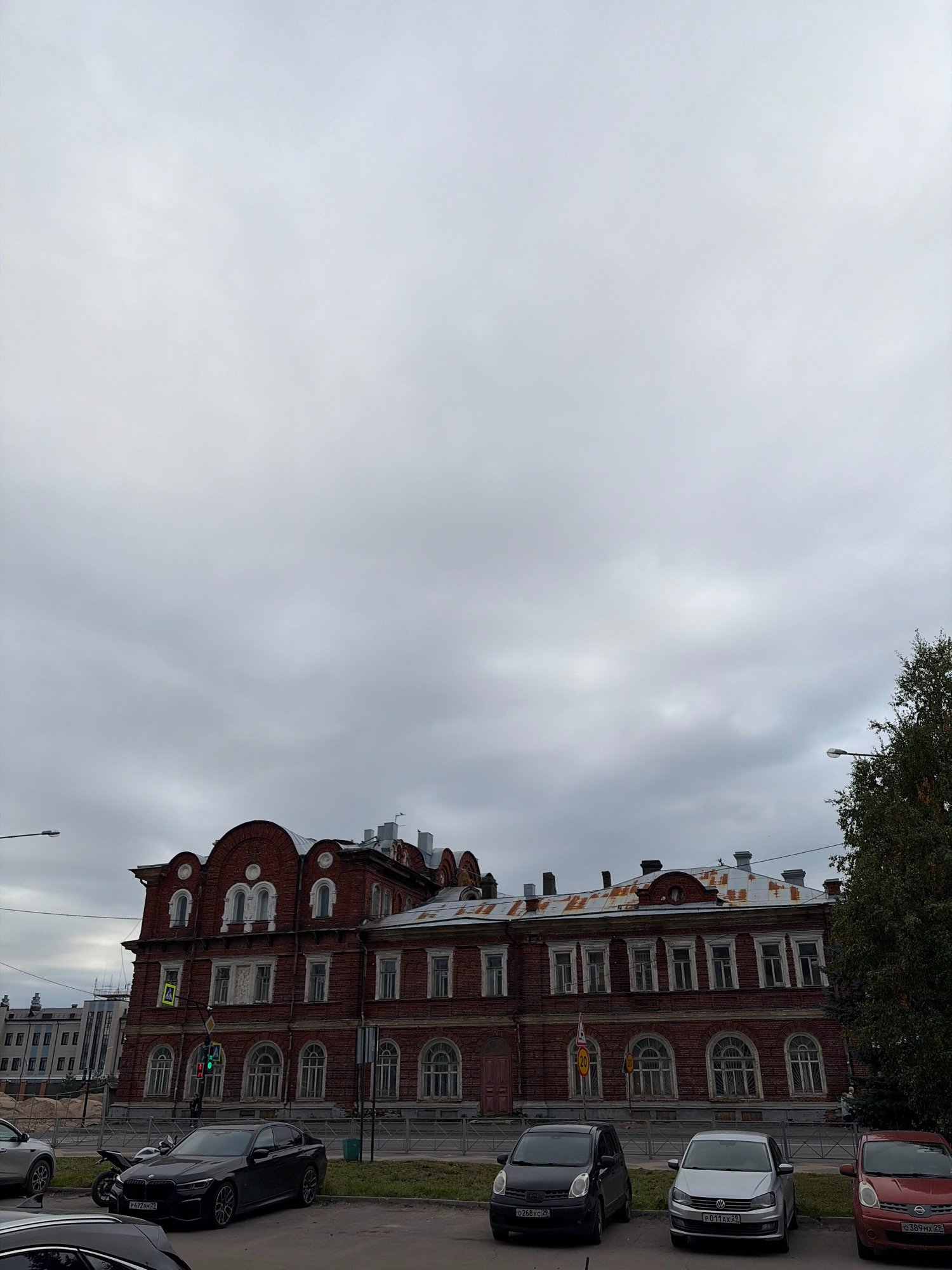 This image shows a view of the historic building at 18 Vologodskaya Street in Arkhangelsk, Russia, on an overcast day. The red brick building features arched windows with white detailing and a weathered, rusty metal roof. In the foreground, several cars are parked, including a black BMW sedan with a license plate ending in "29", a silver Volkswagen hatchback, and a red Nissan hatchback with a license plate ending in "29". A motorcycle is also visible parked near the building. A traffic light, currently showing red and green, is positioned on the street. Further down the street, more modern buildings are visible, and a tall tree with dark green foliage stands to the right of the frame. The sky is filled with heavy, gray clouds, suggesting a cool or damp day. No people are clearly discernible in the scene, though a figure is vaguely visible near the red car.