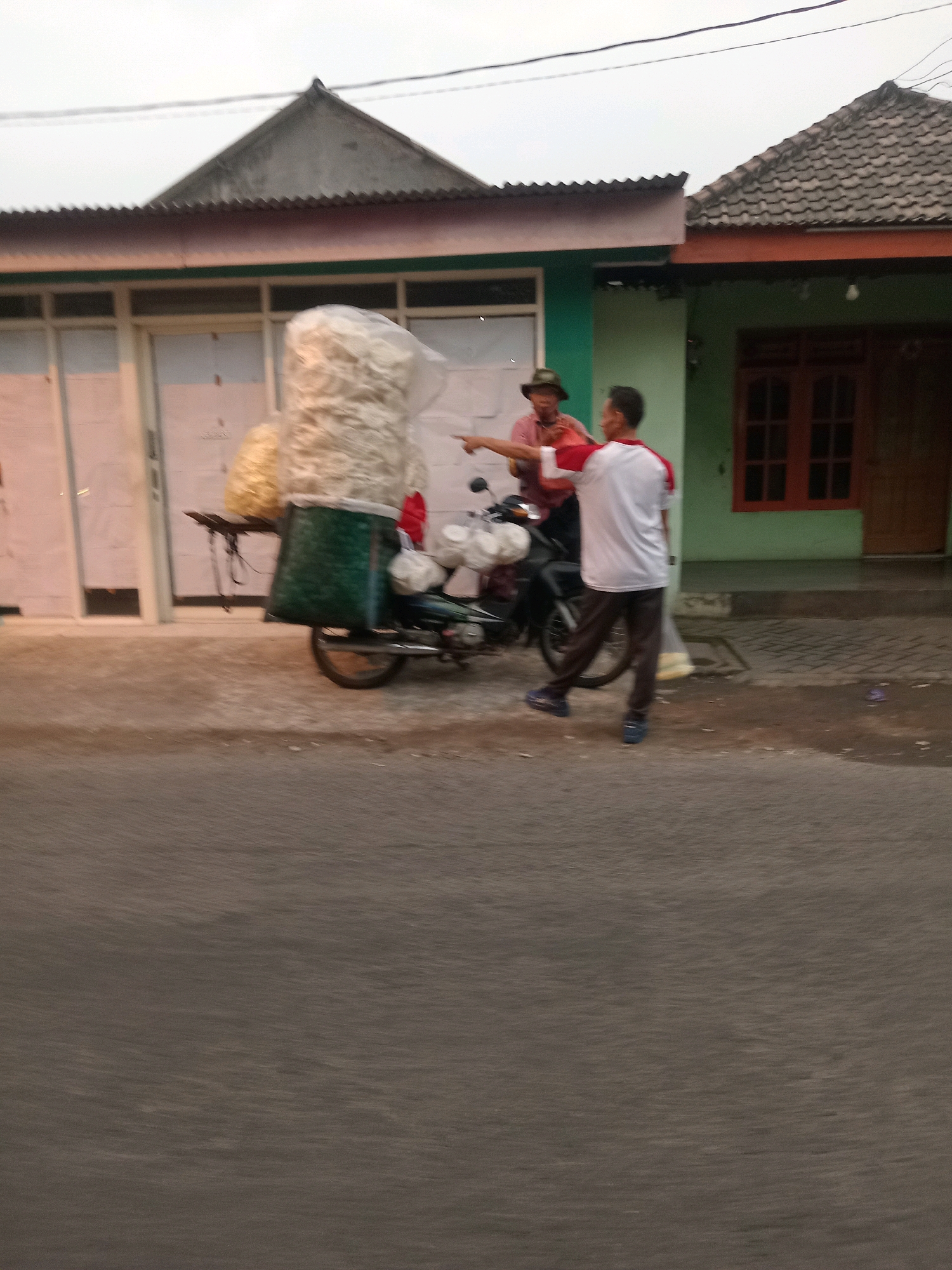 The image depicts an outdoor scene in Kwangsan, Indonesia, near a building with a green facade and a dark-tiled roof. In the foreground, a person wearing a white shirt with red accents and dark pants walks away from a parked motorcycle. The motorcycle is loaded with large bags of what appears to be food products, possibly snacks or chips, covered in plastic. To the left of the motorcycle, another individual, wearing a hat and a red shirt, is gesturing towards the person in the white shirt. The interaction suggests a transaction or conversation related to the goods on the motorcycle. The environment appears to be a street or sidewalk in front of a residential or commercial area, with a cloudy sky overhead. The image is slightly blurred, indicating motion or a low-resolution capture.