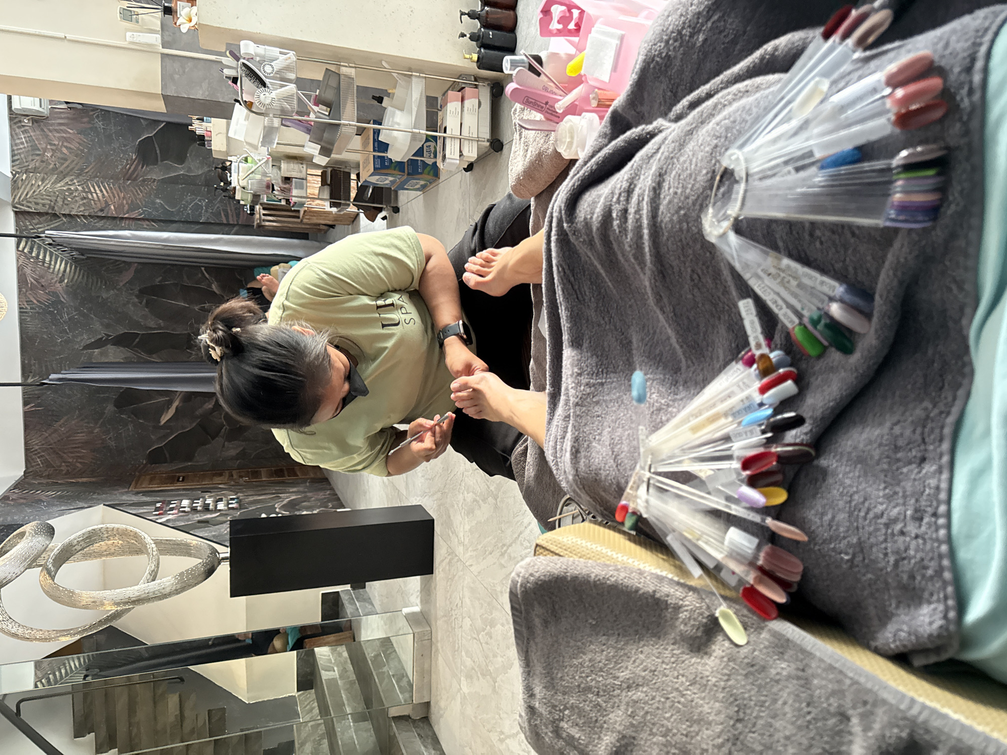 In Denpasar, Indonesia, a person is receiving a pedicure at a nail salon. The client is lying down, covered by a grey blanket, with their feet outstretched. A nail technician, wearing a light green t-shirt and a face mask, is kneeling and working on the client's toes with a tool. Various nail polish color samples are fanned out on the blanket near the client's feet, indicating a selection process. The salon's interior features a modern design with patterned wallpaper, a staircase with mirrored railings, and shelves stocked with nail care products.