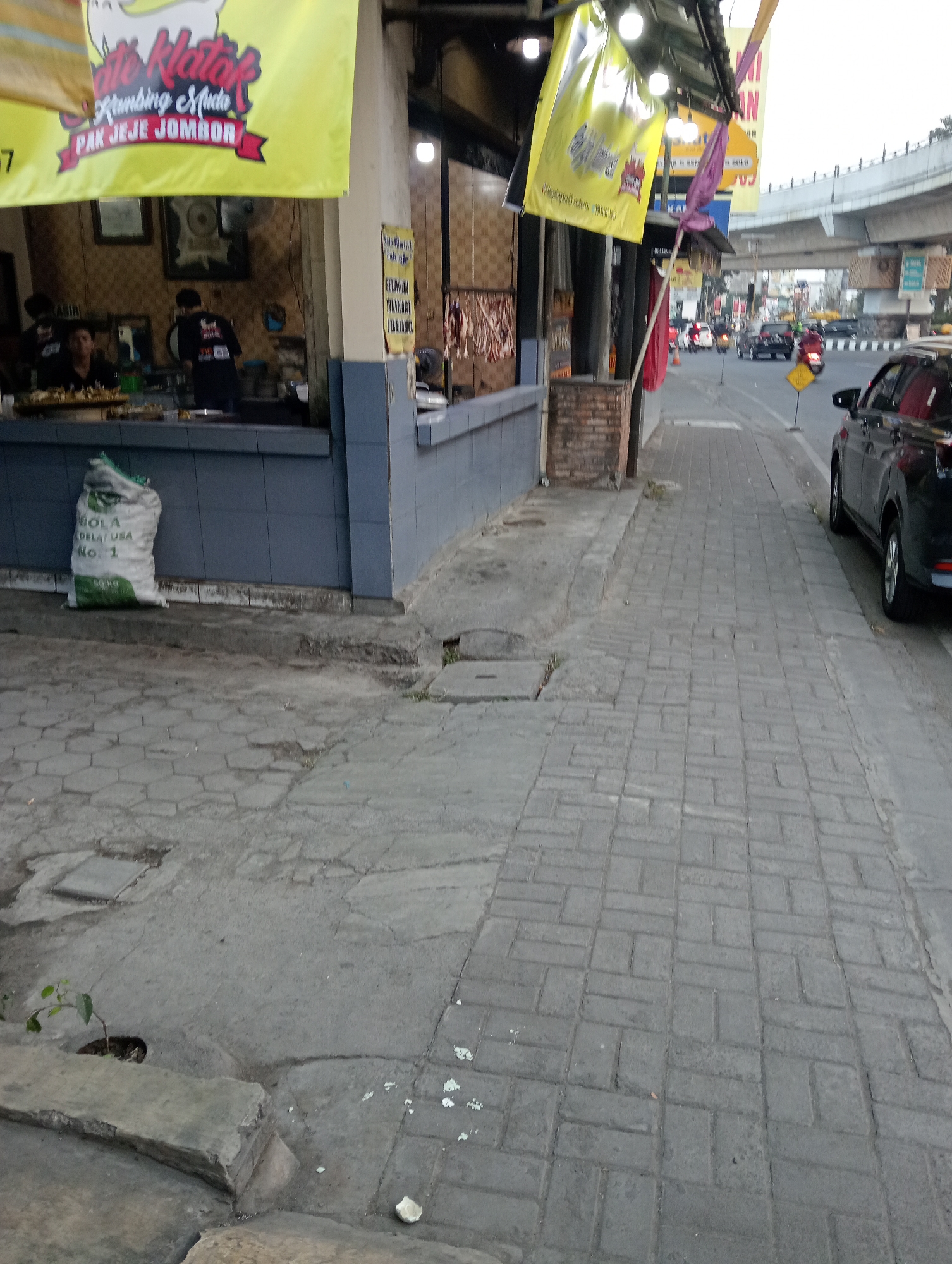 The image depicts a street scene in Sinduadi, Indonesia, at eye level. A food stall is visible on the left, with yellow banners displaying Indonesian text. One banner reads "Ate Klotok Kambing Muda Pak Jeje Jombor". Inside the stall, two people are seen preparing food. In front of the stall, a white sack labeled "BOLA DELAT USA NO. 1" is placed on the ground.
The foreground shows a paved sidewalk made of interlocking gray bricks. To the right of the sidewalk, a black SUV is parked. The street continues into the background, with several cars and motorcycles on the road. An elevated highway structure is visible in the distance, spanning across the street. The overall environment suggests a commercial area with moderate traffic.