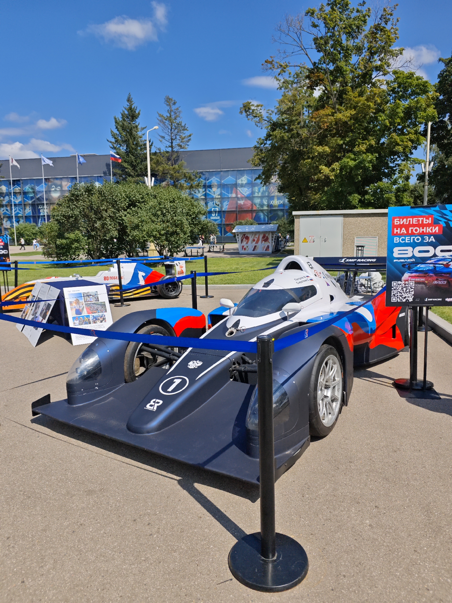 The image depicts an outdoor exhibition of racing cars under a clear blue sky with scattered clouds. The primary subject is a dark blue and white racing car with red and blue accents, bearing the number "1" and the SMP Racing logo. It is positioned in the foreground, cordoned off by blue velvet ropes and black stanchions. Behind this car, another white racing car with orange and blue detailing is partially visible. To the right, a large blue poster advertises "Tickets for races for only 800 rubles" and includes a QR code. In the background, there is a large building with a patterned glass facade, likely a sports or exhibition venue. Trees with green foliage are visible, and several people are walking in the distance. The ground is paved with asphalt. The presence of the racing cars and the event-related poster suggests this scene is likely taking place at a motorsport-related venue or event, possibly within a city park or exhibition area in Moscow, Russia, given the mention of "SMP Racing" which is a Russian motorsport program.