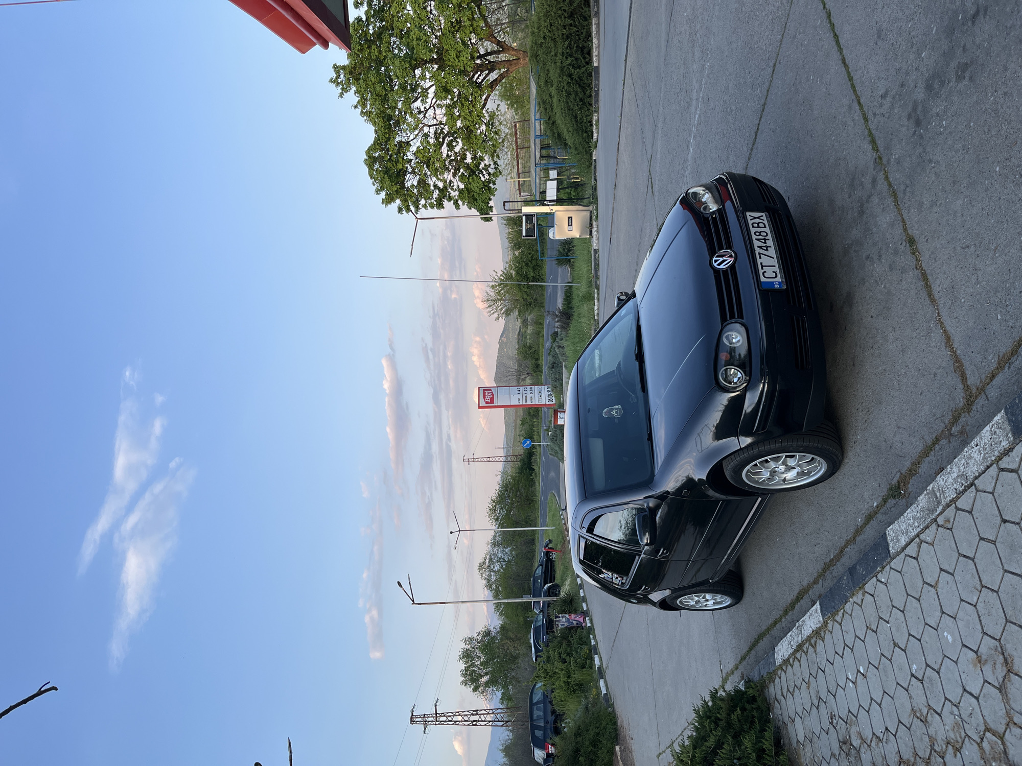 The image depicts a black Volkswagen Golf parked near a gas station in Mihiltsi, Bulgaria. The car's license plate number is CT 7448 BX. The gas station has red and white signage with visible price postings. The surrounding environment includes trees, other parked cars, a telephone pole, and a mountain range visible in the background under a partly cloudy sky.