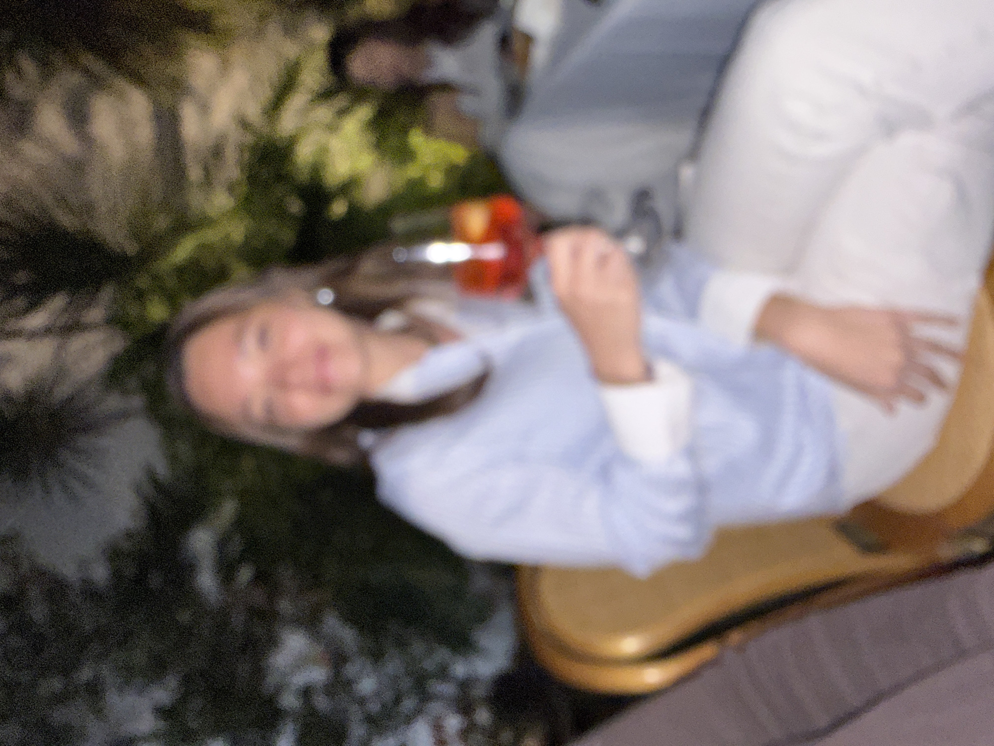 The blurry image displays a low-light scene, likely an outdoor or patio setting within the city of Puebla, Mexico. A female individual is centrally positioned, seated and facing forward, exhibiting a faint smile. She wears a light blue long-sleeved collared shirt with prominent white cuffs and white pants. Her right hand holds a stemmed glass containing a vibrant red-orange liquid, indicative of a cocktail. Her hair is dark, and skin tone appears fair. The subject is seated on a chair, the upper left portion of which, featuring a brown-gold upholstered backrest, is visible. The background consists of heavily out-of-focus dark green foliage, consistent with trees or shrubs, marked by scattered areas of artificial illumination. A second, vaguely discernible individual, likely female, is seated in the upper right background, also out of focus and partially obscured by the foreground subject.