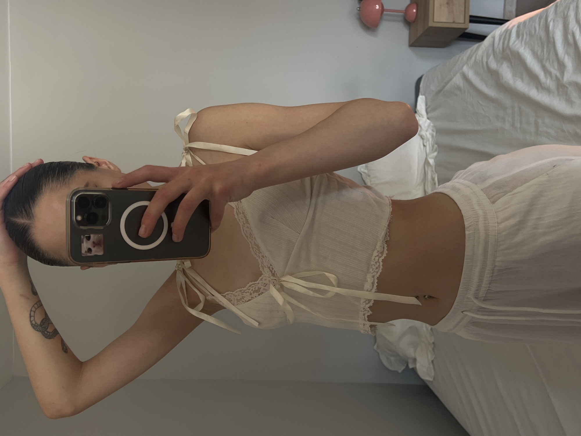 A young woman is taking a mirror selfie in a bedroom. She is wearing a white tie-strap crop top with lace trim and white drawstring pants. She has a snake tattoo on her left arm. The room has a bed with white bedding, a nightstand with a pink lamp, and a wall. The woman's face is obscured by her phone. The image is taken from the waist up. There is no information to indicate this is Ternopil, Ukraine.