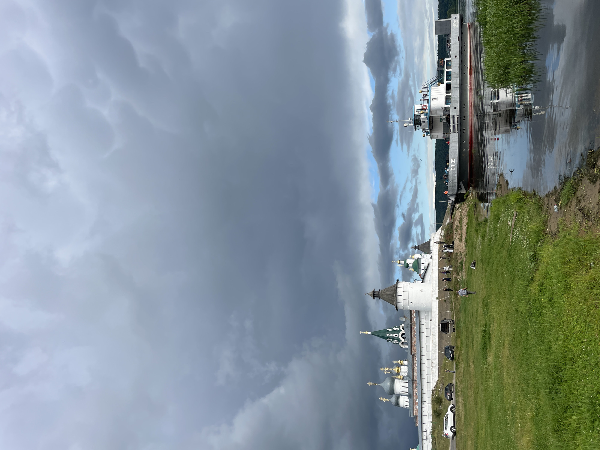 In Makar'evo, Russia, a cargo ferry, labeled "СП-35," navigates a body of water. The ferry, a gray and white vessel with red accents, is positioned near the shore, where tall green reeds grow. On the opposite bank, a historic complex featuring white walls and multiple domed churches with golden crosses stands against a dramatic, cloudy sky. Several cars are parked along the road next to the complex, and a few individuals are visible on the grounds. The water reflects the sky and the ferry, creating a mirrored image.