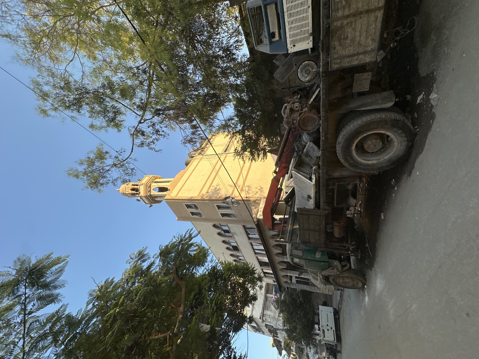 A low-angle, eye-level shot captures a street scene in an unknown city. In the foreground, a large, old green truck with a flatbed is parked on the right side of the street. The flatbed is loaded with various mechanical parts and debris, including what appears to be an engine and other metallic components. A red crane arm is attached to the truck. To the right of the truck, a portion of a white and blue vehicle's cab is visible, also loaded with debris. The street itself appears to be paved and shows some scattered white debris.
Behind the truck, a multi-story building with a mosque-like minaret on top is prominently featured. The building is beige and has arched windows on the upper floors. The minaret is tall and has a decorative top. Sunlight illuminates the building, casting shadows. To the left of this building, another, taller, lighter-colored building is visible, with multiple windows. Trees with green foliage frame the scene from the left and right, with some branches extending into the frame. Power lines are visible crisscrossing the sky. In the background, to the far left, a white generator is parked, with a person standing nearby. The sky is clear and blue, suggesting a sunny day.
