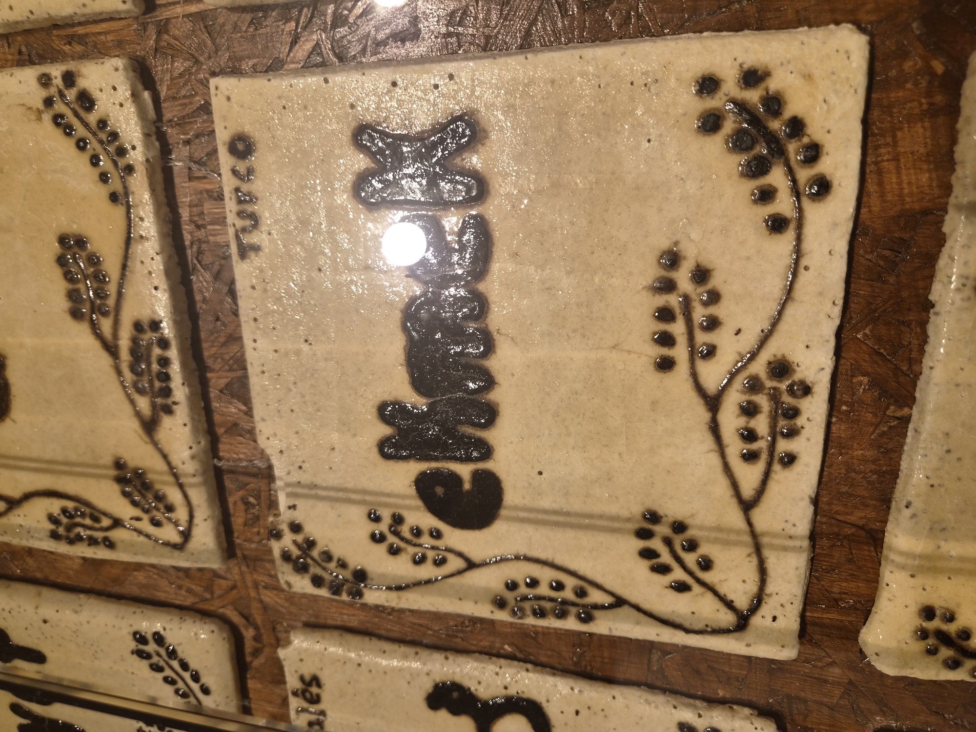 The image shows several ceramic tiles, each with a decorative pattern and some with lettering. One tile in the center has the word "chmeck" written in raised black lettering. To the top right of this tile, another tile has the word "TURCO" in raised black lettering. The tiles are laid on a wooden surface. This display of ceramic tiles is located in Seia, Portugal.