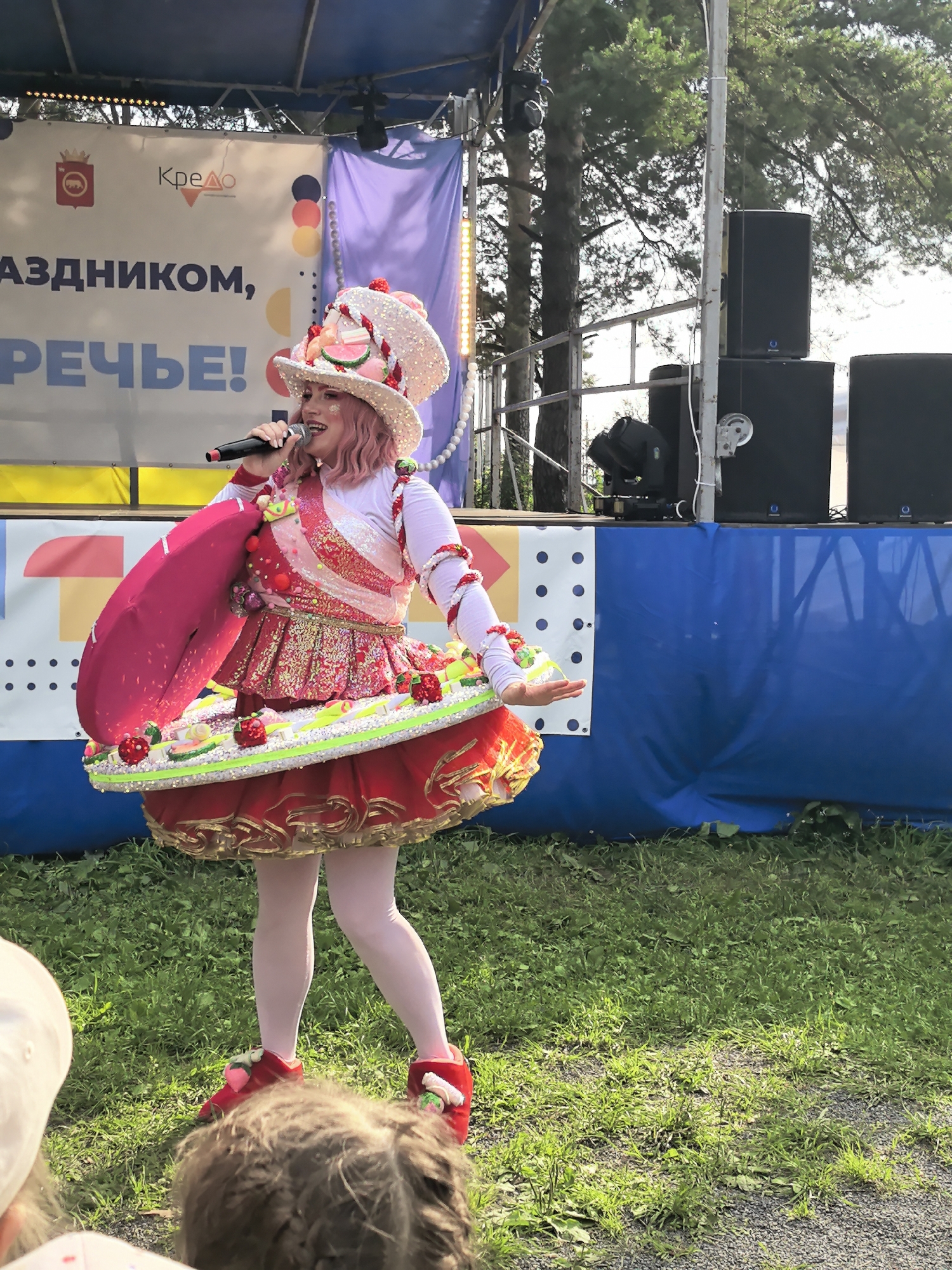 A woman dressed in a brightly colored, elaborate costume, resembling a cupcake or cake, stands on a grassy outdoor stage. She has pink hair and is holding a microphone, appearing to be performing or speaking. Her costume features a large, decorated hat, a sequined bodice, and a voluminous skirt adorned with fruit decorations. Behind her, a banner with Russian text and logos is visible, as well as speakers and trees. The background suggests a public event or festival. The image was taken in the city of Gorniy, Russia.