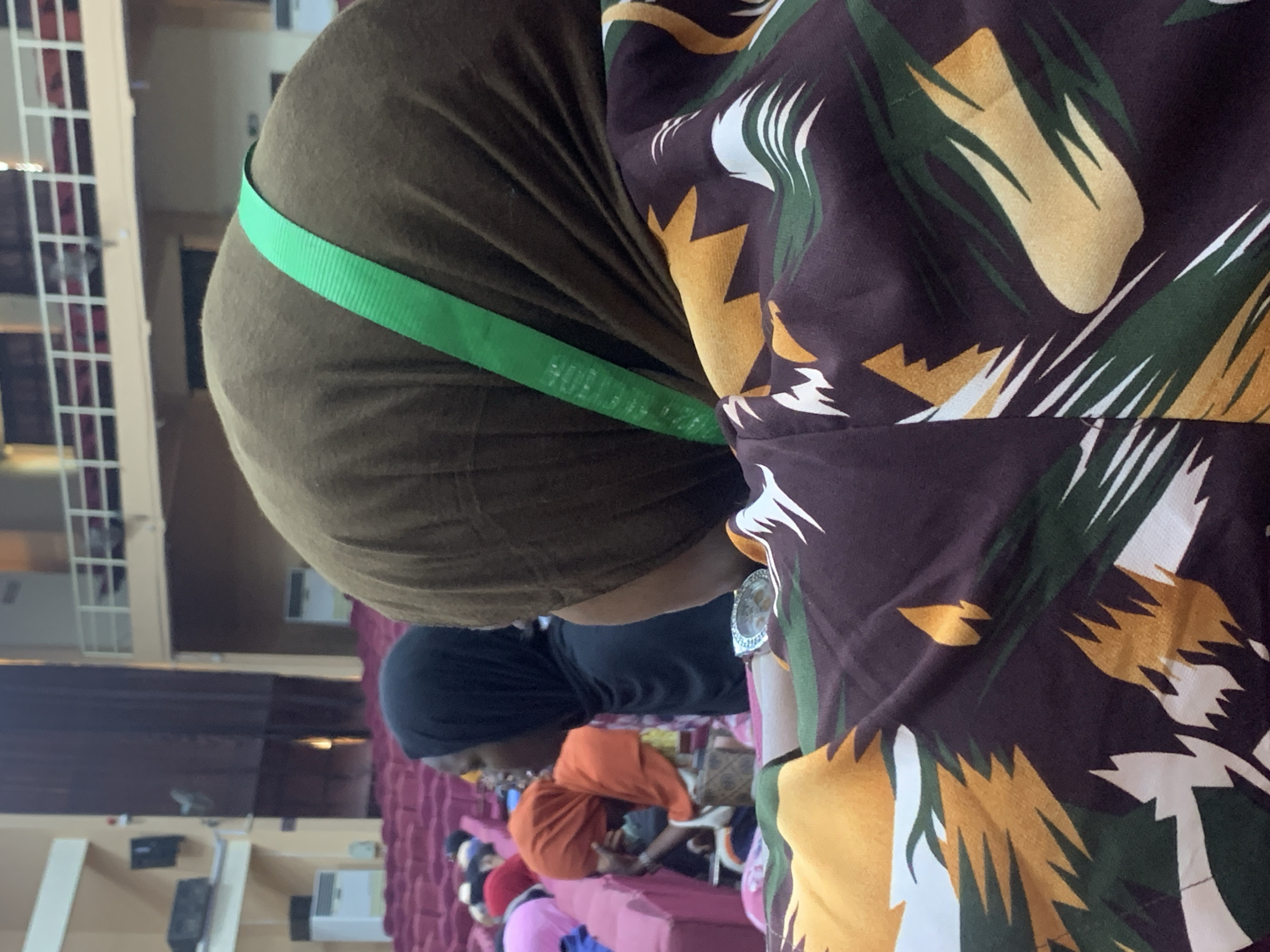 The image captures an indoor gathering or event, seen from an over-the-shoulder perspective of a person in the foreground. The scene is rotated 90 degrees counter-clockwise.
In the immediate foreground, the back and right shoulder of an individual wearing a dark brown hijab or head covering are visible. They are also wearing a patterned garment with a dominant dark brown background, featuring abstract designs in yellow, dark green, and white, resembling stylized foliage or brushstrokes. A bright green ribbed strap, possibly a lanyard or bag strap, crosses their right shoulder and back. Faint text is visible on the green strap, including "VICTOR" and what appears to be "UNIVERSITY OF ILORIN", suggesting a connection to the institution in Ilorin, Nigeria.
Beyond the foreground subject, several other individuals are present in the midground and background, most of whom are seated. One person with a black head covering and dark top is visible. Further back, another person wearing an orange head covering can be seen.
The setting is a large indoor space, resembling an auditorium, lecture hall, or multi-purpose event room. Rows of red or maroon upholstered seats are visible in the background, suggesting an audience setup. Light brown or beige walls form the backdrop, with some white rectangular structures, possibly railings of a balcony or upper level, on the left side of the frame. Several black boxes, likely speakers, and white wall-mounted units, possibly air conditioners or electrical panels, are affixed to the walls. The overall impression is that of a formal or semi-formal gathering taking place in a public or institutional building in Ilorin, Nigeria.