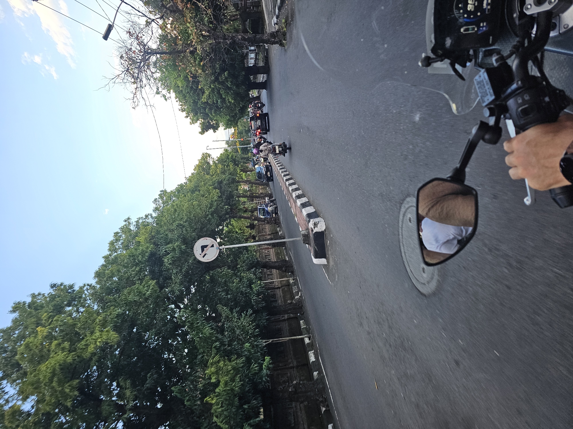 Captured from the vantage point of a moving motorcycle, the image presents a street in Denpasar, Indonesia. The perspective is looking forward, down the road. The rider's left hand is visible, gripping the handlebar and controls. A side mirror reflects a glimpse of the rider. The road extends into the distance, lined with trees on the left side. A "no left turn" traffic sign is mounted along the side. Several vehicles, including other motorcycles and cars, are visible further down the street. The sky is light blue with some clouds. The overall light suggests it is daytime.