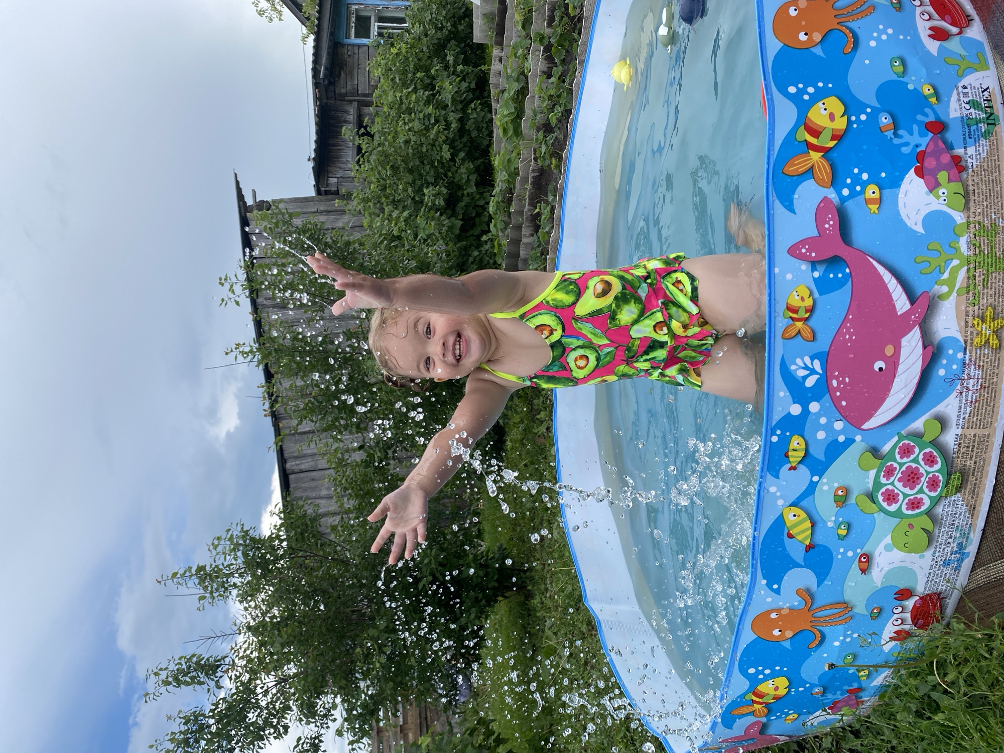 A young girl with blonde hair, wearing a pink swimsuit with an avocado pattern, is splashing in a small, inflatable pool. She is leaning back with her arms outstretched, and water is spraying around her. The pool has a colorful design featuring marine life like a whale, octopus, crab, and turtle.
The scene is outdoors, with lush green trees and bushes in the background. A rustic wooden building, possibly a house or shed, is visible behind the pool, suggesting a rural or backyard setting. The sky is overcast, indicating a cloudy day. The girl appears joyful and excited, her mouth open in a wide smile as she plays in the water.
The text on the pool's edge is a warning in multiple languages, including Russian, stating "WARNING! Drowning hazard. Never leave children unattended." Other visible text includes "INTEX" and "Made in China." The overall impression is one of playful summer fun in a simple, outdoor environment.