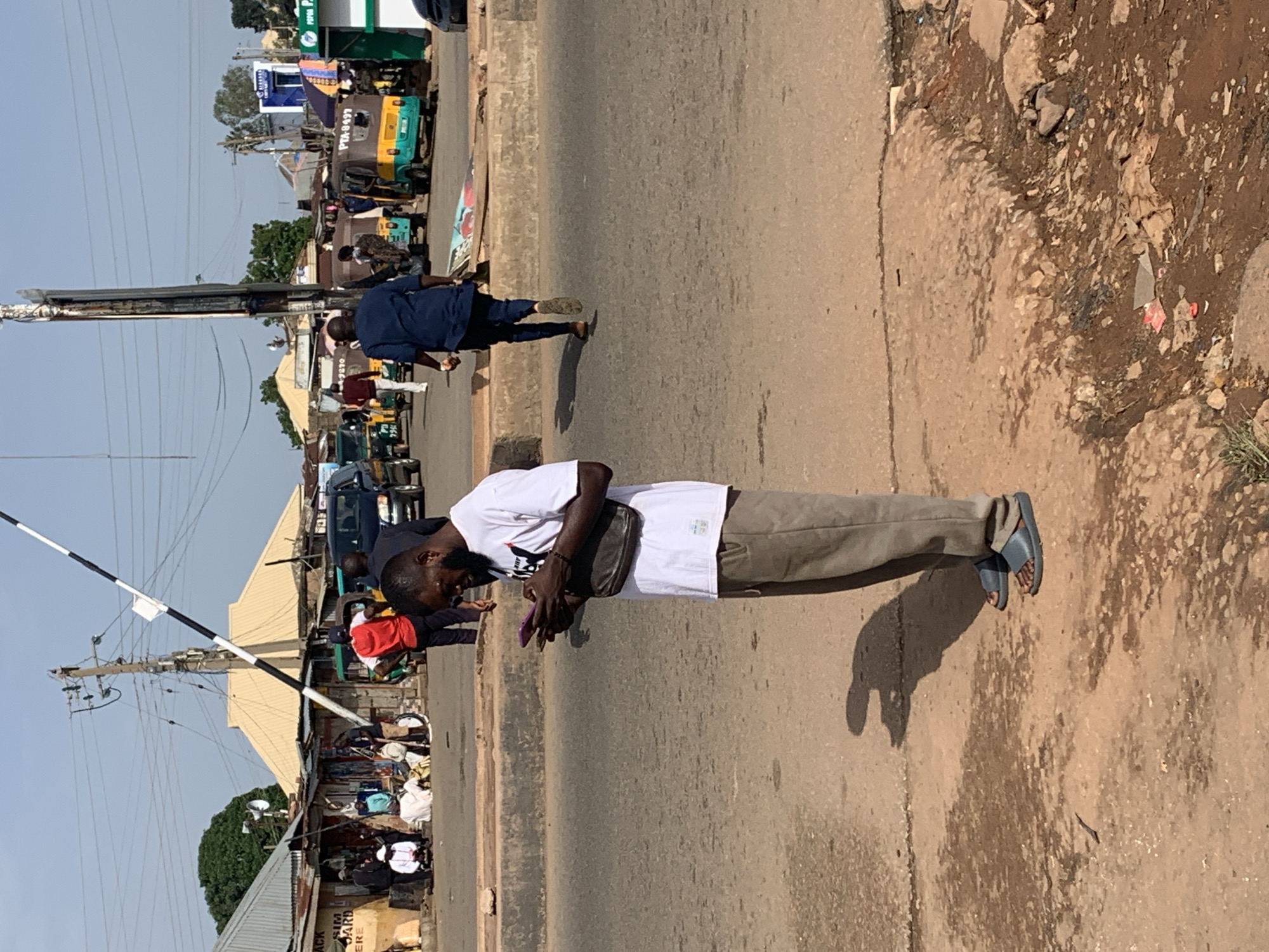 This image depicts a street scene in Gura Top, Nigeria, under a clear sky. The foreground shows a man wearing a white t-shirt and grey trousers, standing on a dirt and asphalt surface, engrossed in his smartphone. He has a dark crossbody bag slung over his shoulder. Behind him, a paved road is visible, with a concrete barrier separating the road from the pedestrian area. A dark-colored SUV is parked on the far side of the road, along with several open-air market stalls and people milling about. A yellow and green auto-rickshaw is also present. A black and white striped barrier is raised, suggesting it might be a checkpoint or an entrance. The background features more buildings, some with corrugated metal roofs, and a prominent utility pole with electrical wires. The overall environment appears to be a busy urban setting with signs of commerce and everyday life.