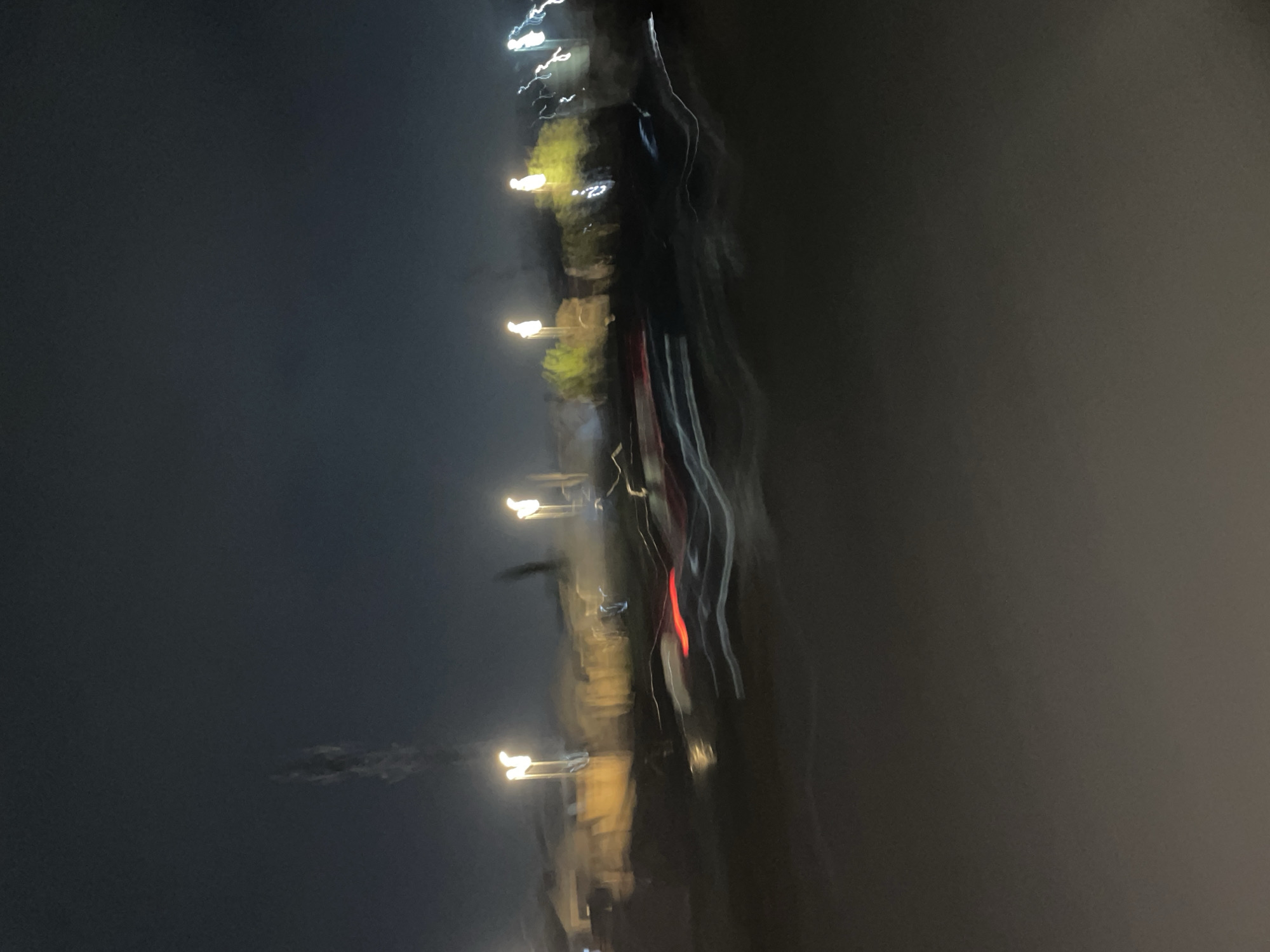 This abstract image captures a nocturnal scene characterized by pronounced motion blur and light streaks. The dominant feature is a vertical array of elongated, bright lights, primarily in warm yellow-orange and white hues, appearing against a dark, indistinct background. A distinct vertical red light streak is visible towards the right side of the image, alongside other brighter white light trails.
These light streaks suggest artificial illumination, possibly from streetlights, vehicle lights, or illuminated buildings, captured with either a moving camera or during a long exposure. A blurred, dark, vertical mass extends through the central and right portions of the frame, which seems to be the source or backdrop for some of these lights, potentially representing buildings, trees, or other structures along a path or road.
The setting is clearly nighttime, as indicated by the deep dark tones and the reliance on artificial light sources. The heavy blurring creates a dynamic, almost painterly effect, implying speed or continuous movement. The atmosphere appears hazy or indistinct, an effect enhanced by the blur. There are no identifiable people, specific objects, or visible text within the frame. The overall impression is one of movement and light in an urban or developed environment. The location context is Abuja, Nigeria.