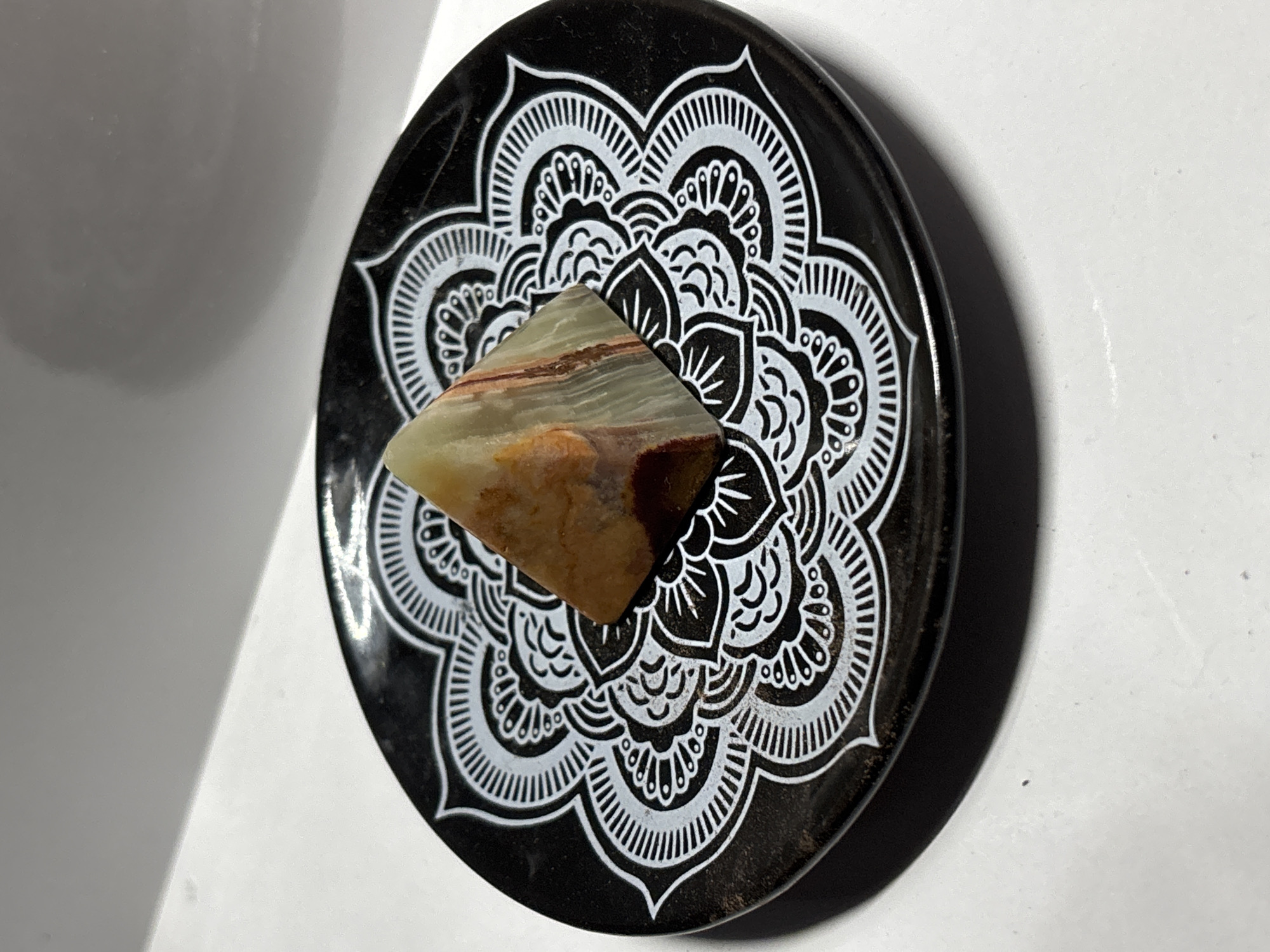 A small, polished, multi-colored gemstone pyramid, exhibiting bands of green, white, and reddish-brown, rests on a black, circular dish adorned with a white, intricate mandala-like design. The dish is positioned on a white surface, with a subtle shadow cast by the object and dish. The background is a plain, white, wall-like structure that angles upwards. The image is brightly lit, with a slight glare visible on the dish and the white surface. The scene depicts a still life arrangement of decorative objects. There is no discernible location in Gurkovo, Bulgaria.