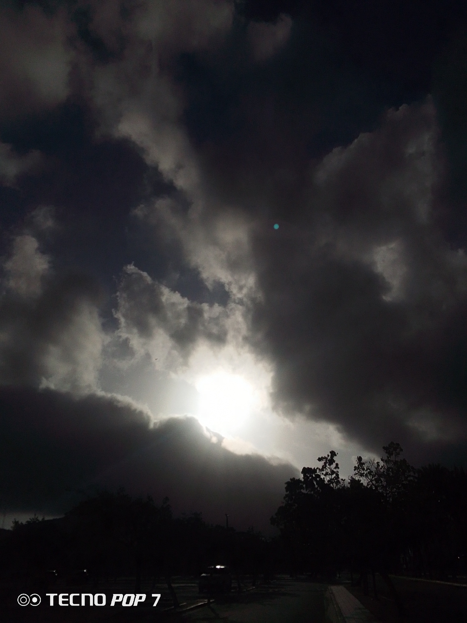 The photograph captures a dramatic sky in Pedro González, Venezuela, dominated by heavy, dark clouds obscuring the sun. A bright light source is present behind the clouds. The landscape below appears dark and silhouetted, showing trees and possibly some structures. The overall scene conveys a sense of impending weather or a darkening evening. The "Tecno Pop 7" branding suggests it was taken using a mobile phone camera.