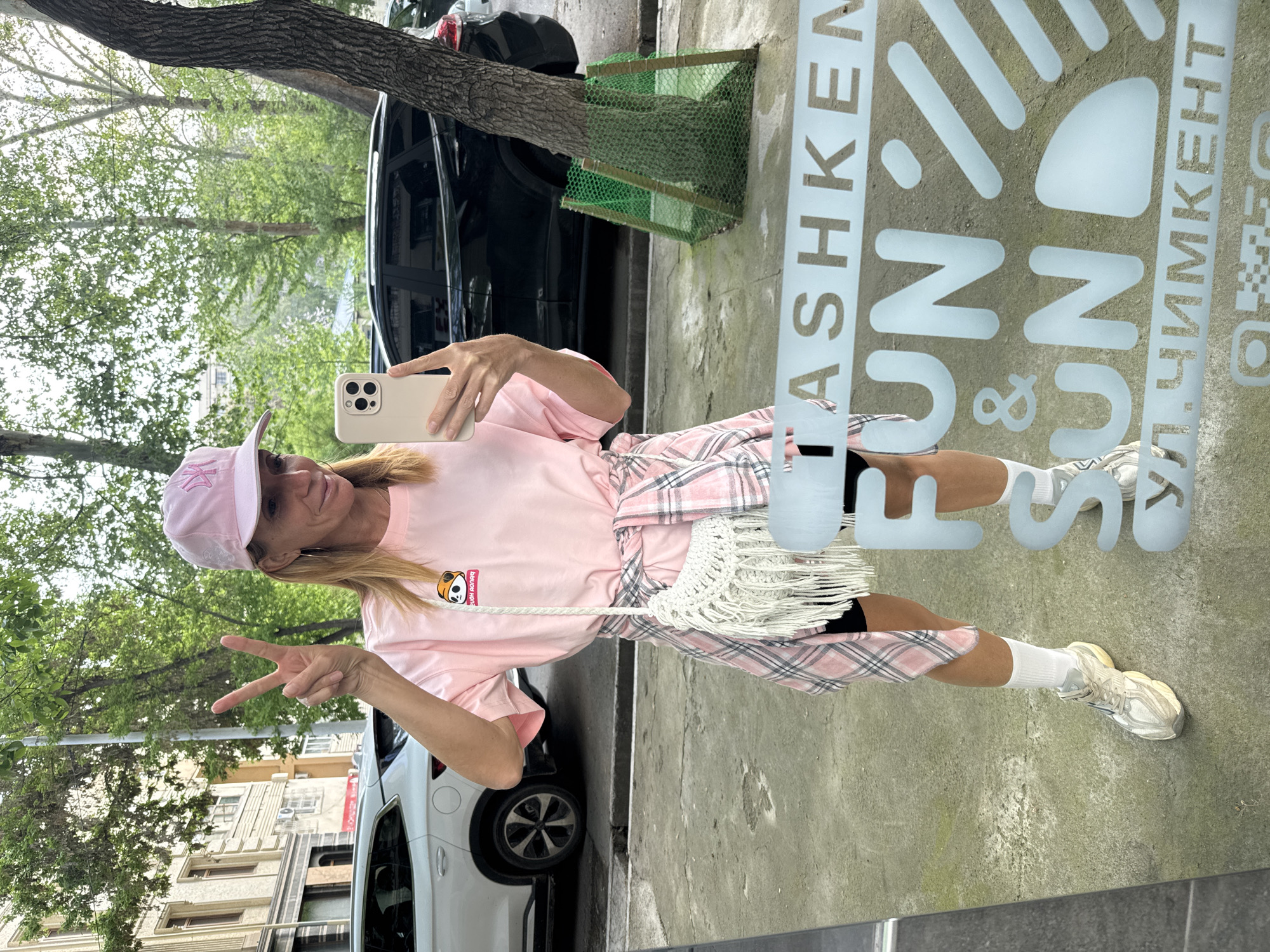 A woman in a pink outfit stands in front of a glass window, taking a selfie with her smartphone. She wears a pink T-shirt, a pink plaid skirt, white socks, and sneakers. She is also wearing a pink baseball cap and has a white tasseled bag slung over her shoulder. With her left hand, she makes a peace sign. The window has the words "TASHKENT FUN & FUN" written on it in white letters. The background includes city elements like trees, buildings, and cars. The location appears to be in Tashkent, Uzbekistan, as indicated by the text on the window.