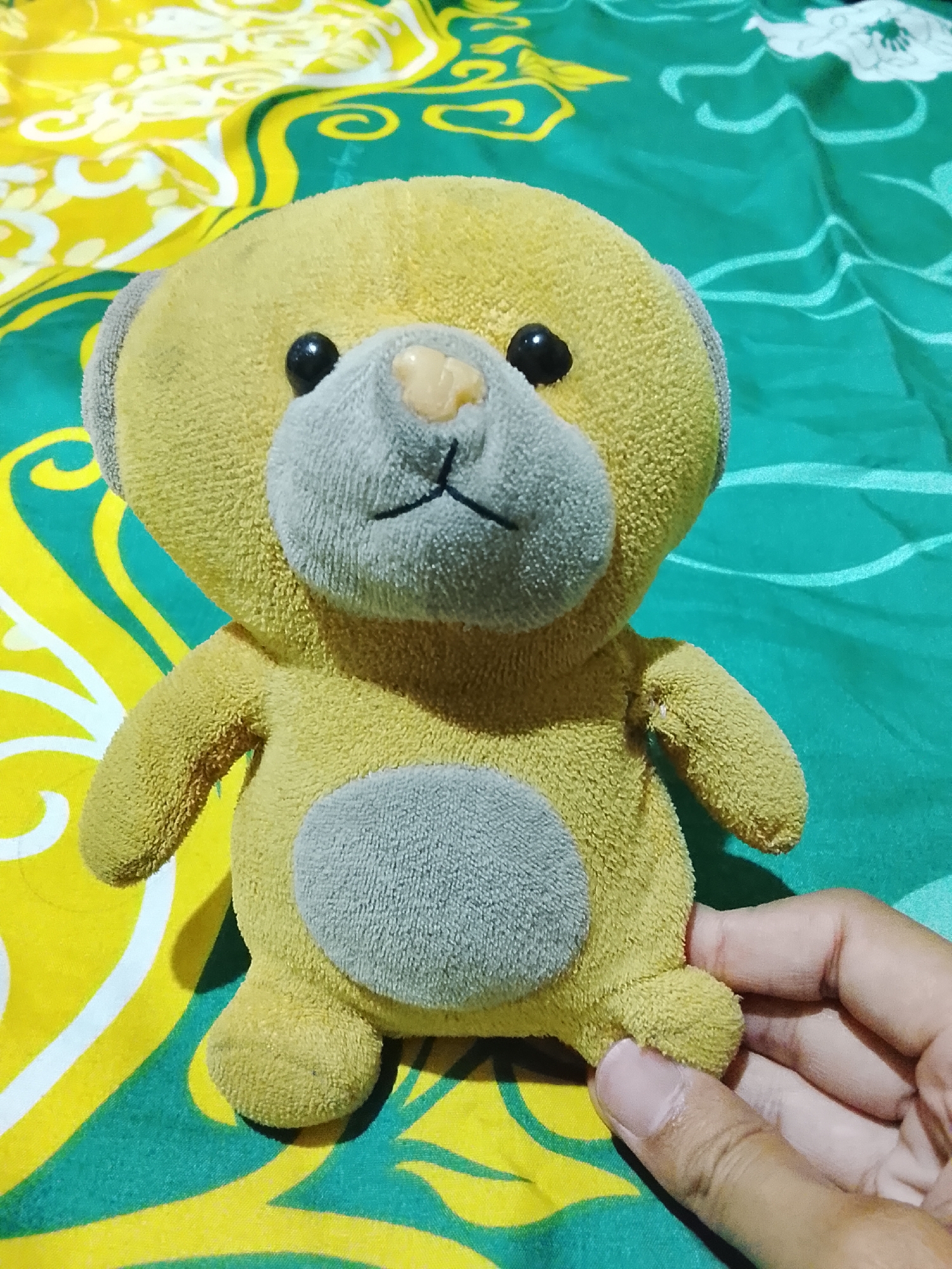 A yellow plush teddy bear with gray inner ears, muzzle, and oval belly sits on a green and yellow patterned sheet. The teddy bear's eyes are black and round, and its nose is a small, light brown nub. Its mouth is represented by two black embroidered lines. A human hand, with visible fingernails, is gently holding the teddy bear's left foot. The sheet has a repeating wavy pattern of green and yellow. The scene appears to be indoors, possibly in a bedroom, given the bedding.