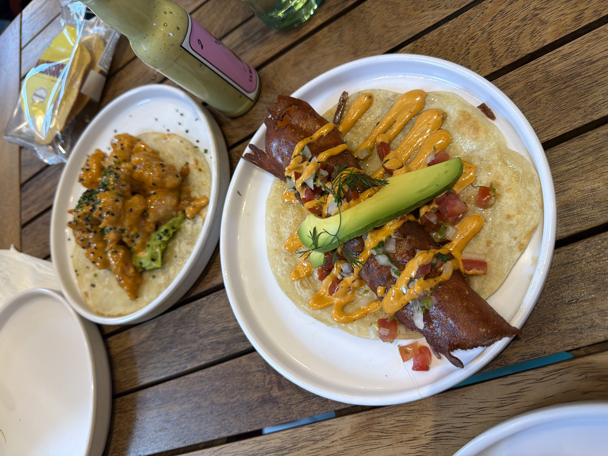 The image depicts two tacos on white plates, arranged on a wooden table. One taco features a large, rectangular piece of cooked protein, topped with diced tomatoes, onions, avocado slices, dill sprigs, and drizzled with an orange sauce. The other taco is filled with breaded and sauced pieces of protein (likely shrimp or chicken) with broccoli and topped with sesame seeds. Next to the tacos are a bottle of light green sauce with a pink label, a clear plastic bag containing cookies, and other plates. The setting appears to be a restaurant in Mexico City, Mexico, based on the style of the food.