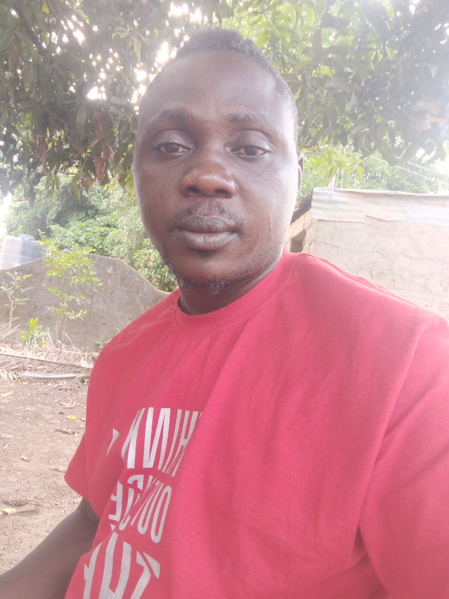 A medium-shot selfie of a man wearing a red t-shirt with white lettering on it. The background shows a dirt ground, a concrete structure, green foliage, a water tank, and what appears to be a partially constructed or dilapidated building. The lighting is bright, possibly suggesting daytime. It's likely in Akwanga, Nigeria, but there are no specific identifiable landmarks.