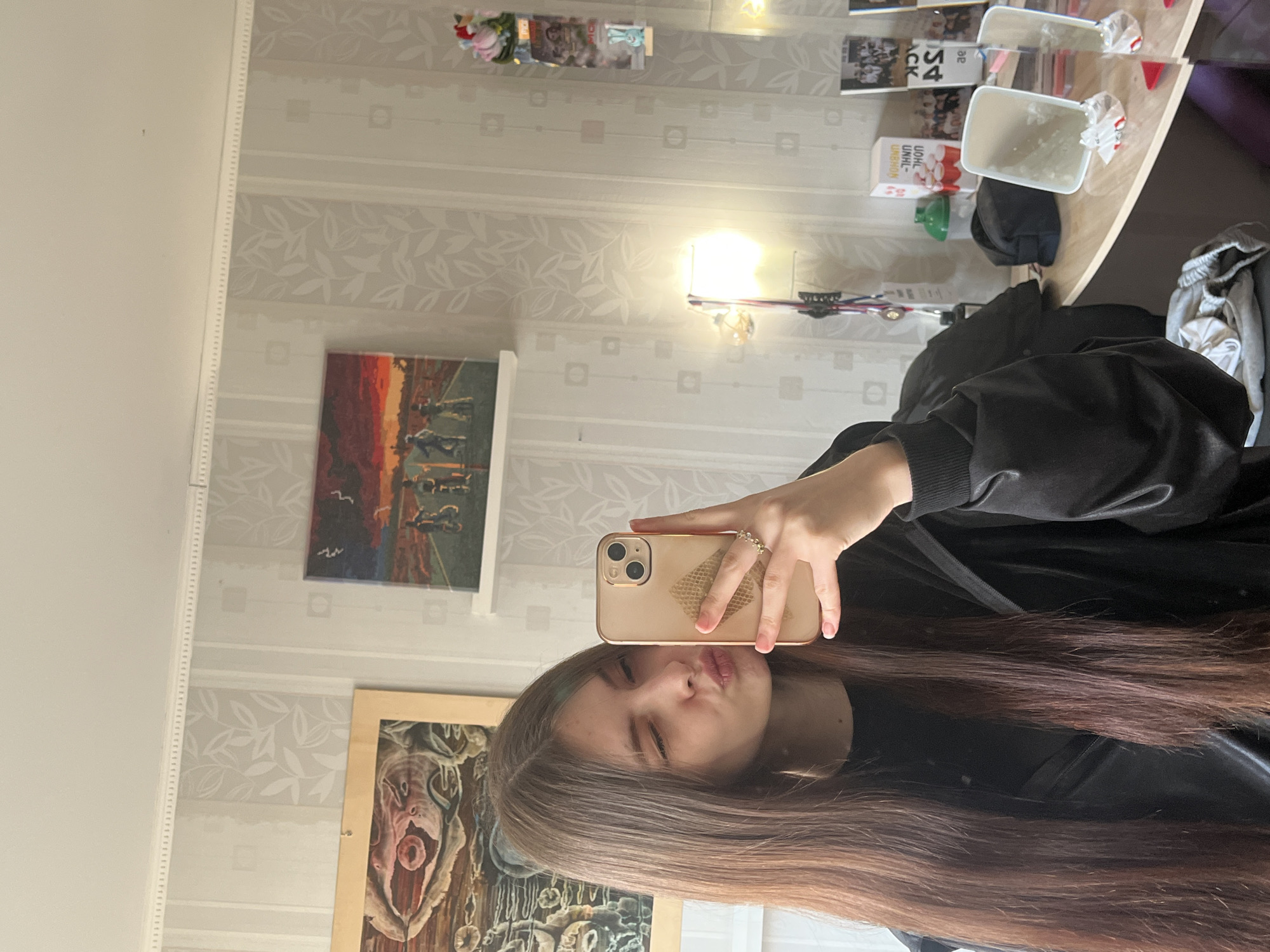 A young woman with long, brown hair takes a selfie in front of a mirror. She is wearing a black top and holding a gold-colored smartphone with a patterned case. Her lips are puckered in a kiss gesture. The background shows a room with light-colored wallpaper with a leaf pattern, various pictures and ornaments on a shelf, paintings on the wall, a lamp, and a table with items including a trash can. The location cannot be reliably determined as Saint Petersburg, Russia based on the information presented in this image.