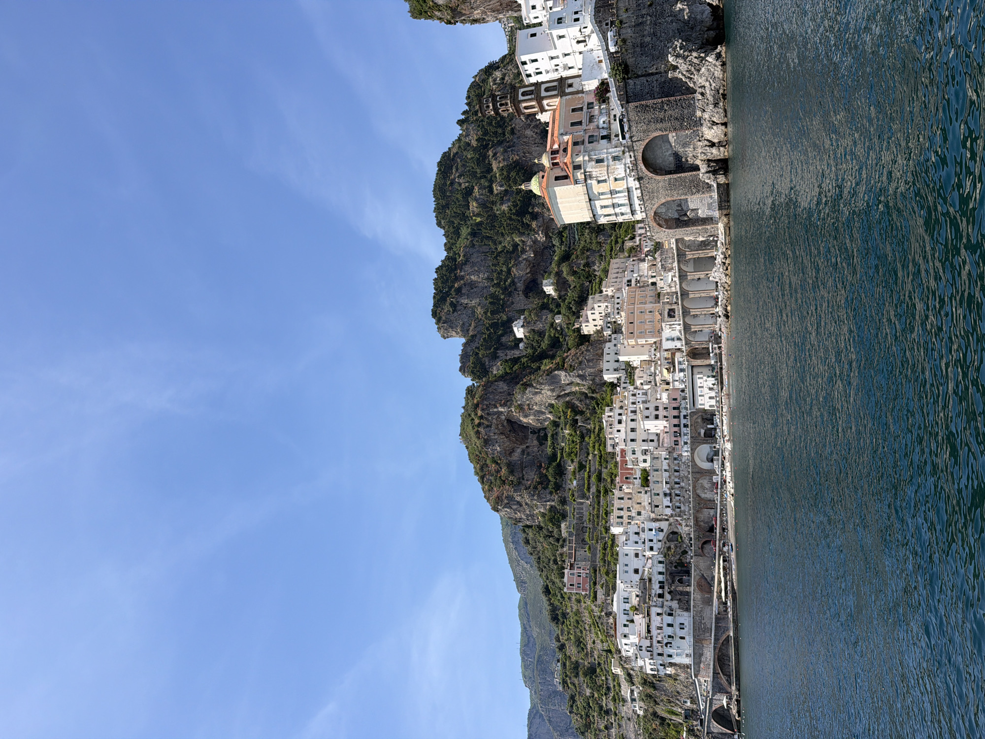 The image depicts a coastal town in Ravello, Italy, situated on a steep cliff overlooking the sea. The town is densely populated with white and pastel-colored buildings. A winding road runs along the coastline. The sky is blue with some wispy clouds. The sea is calm. The buildings are terraced down the cliffside. No people are clearly visible.