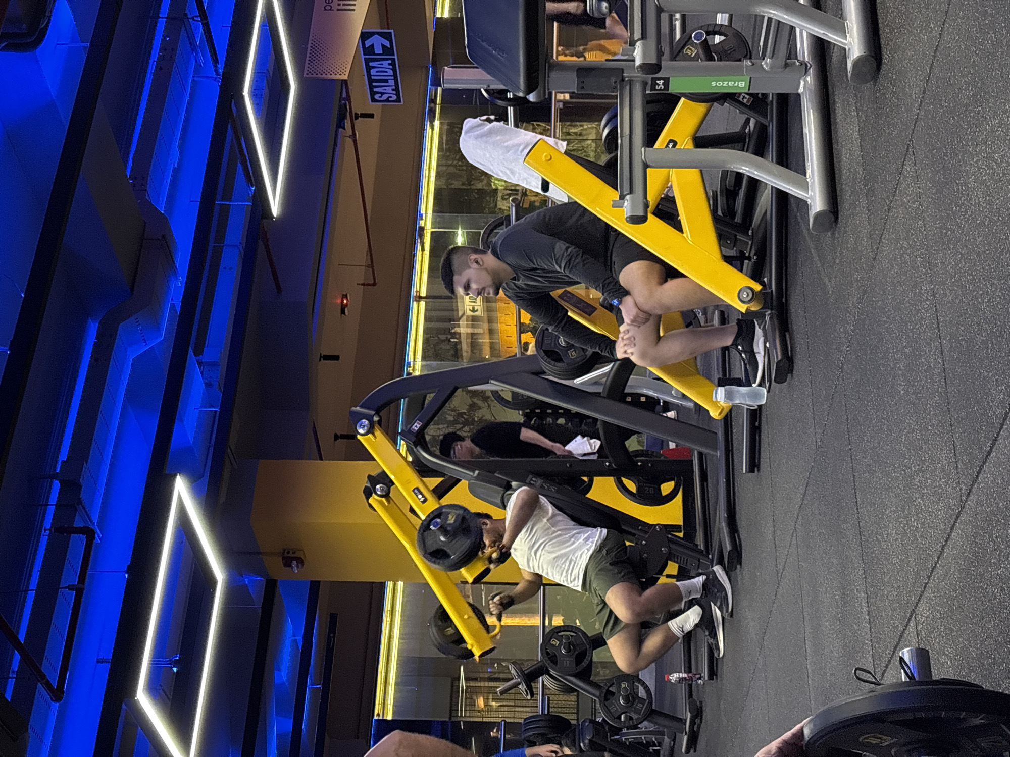 The image depicts an indoor gym, likely within the district of San Isidro, Peru, characterized by its vibrant blue lighting and modern equipment. In the foreground, a man is seated on a yellow weight machine, performing repetitions with a barbell. He is wearing a dark long-sleeved shirt and black shorts. To his left, another man is also seated on a similar machine, engaged in an exercise with a barbell, dressed in a white t-shirt and dark shorts. Several other individuals are present in the background, some exercising and others standing, including a person in a black t-shirt observing the scene. The gym floor is covered with dark rubber matting. Prominent yellow and silver weight machines are visible throughout the space. Above, the ceiling features industrial-style ductwork and lighting fixtures, including bright white rectangular panels. A sign with the word "SALIDA" and an arrow pointing to the right is visible in the upper right portion of the image. The overall ambiance suggests a contemporary fitness facility.