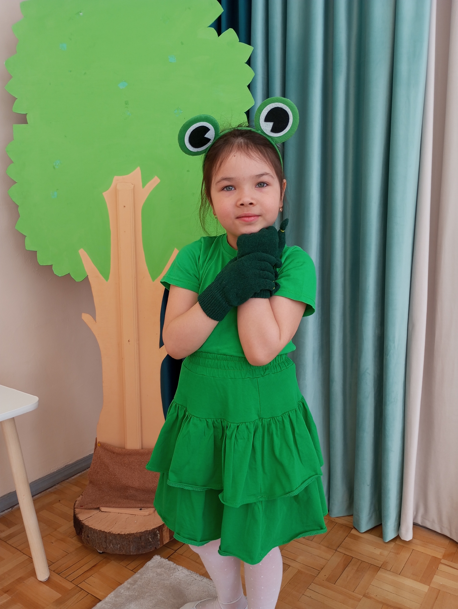 A young girl, likely of primary school age, is depicted in a full-body portrait, dressed in a cheerful green frog costume. The setting is indoors, possibly a classroom or event space, with the provided context indicating Izhevsk, Russia.
The girl wears a bright green short-sleeved t-shirt, a matching tiered green skirt, and dark green gloves. A distinctive green headband with large, round frog eyes, featuring black pupils outlined in white, rests on her head. She also has on white tights adorned with small, light-colored dots. Her hands are clasped together in front of her chest, and she looks directly at the camera with a gentle, somewhat reserved expression. Her eyes are a striking light color, possibly blue or grey, complementing her brown hair.
Behind her, to the left, is a large, flat green cutout of a tree's leafy crown, supported by a light brown wooden cutout of a tree trunk. The trunk stands on a round wooden log slice, forming a stage-like or decorative backdrop. A small portion of a white table with a light wooden leg is visible on the far left. The background on the right features pleated curtains, primarily a light blue/teal hue, with a lighter cream or beige curtain to its right. The floor is covered in light brown wooden parquet, and a small, light-colored rug is seen in the foreground. The scene suggests a themed event, a children's play, or a costume party. There is no visible text in the image.