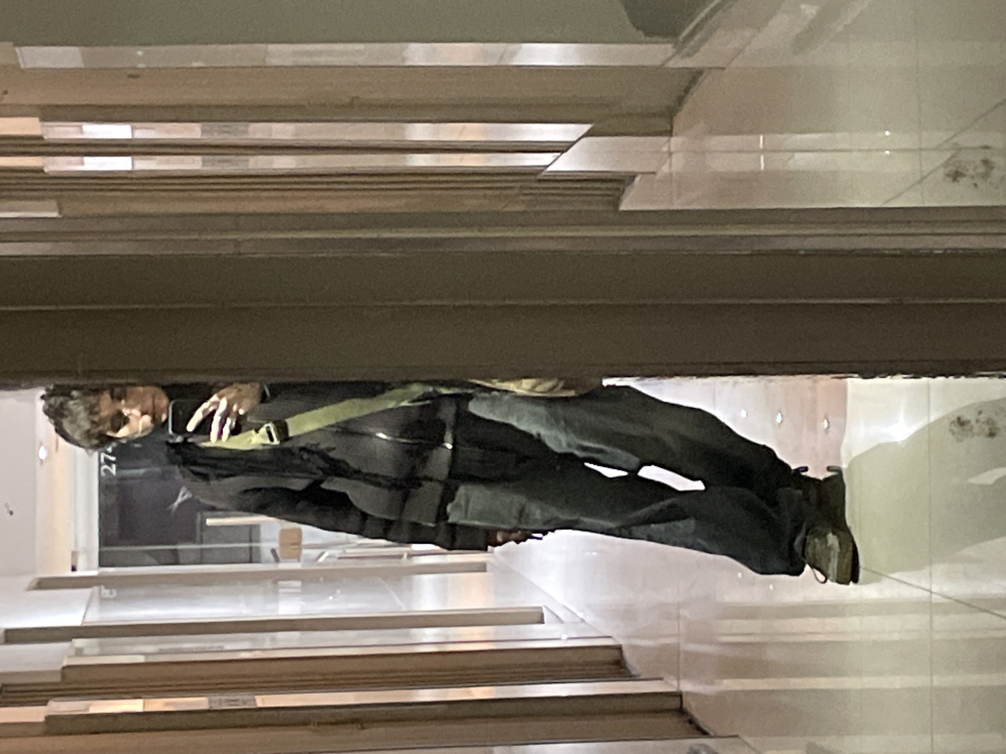 A man stands in front of a storefront window, taking a selfie. The store number "274" is visible in the window. The man is wearing dark clothing, and a shoulder bag is draped across his body. The environment features horizontal lines suggesting tiled steps or shelving. The location is suggested as Buenos Aires, Argentina.