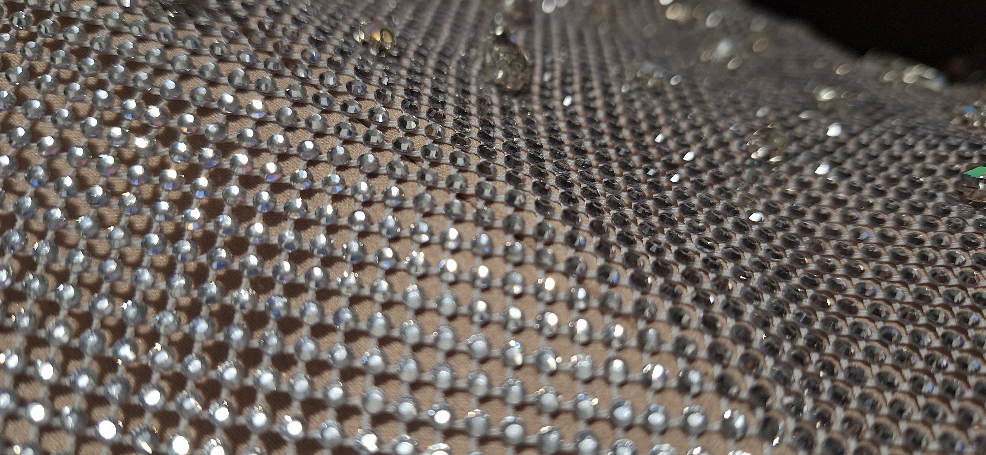 The image shows a close-up of a fabric covered in small, clear rhinestones or beads. The material underneath appears to be a light tan or beige color, providing contrast to the reflective gems. The gems are arranged in a grid-like pattern across the surface. The lighting causes bright reflections on the gems, creating a sparkling effect. The depth of field is shallow, with some areas in sharp focus and others blurred.
There is no information in the image to suggest a location in Blagoevgrad, Bulgaria, or to identify specific people, actions, or interactions. The scene is a purely textural and material-focused close-up.